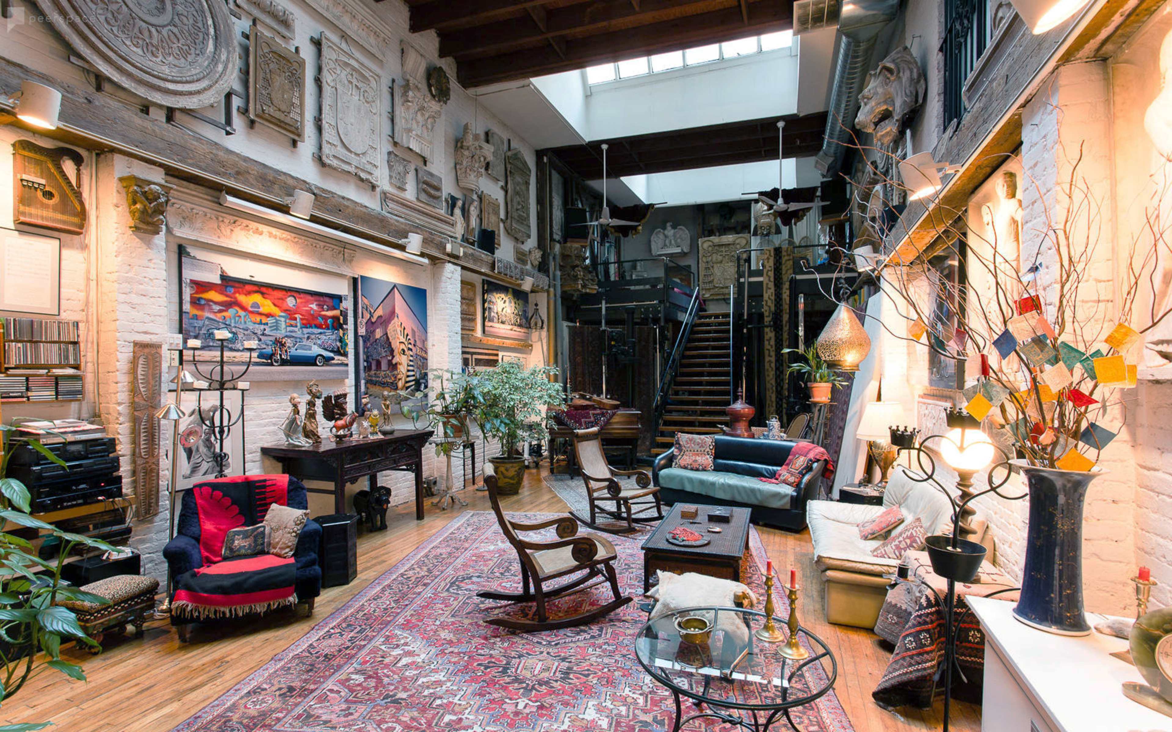 A brightly lit room featuring a mix of seating areas, decorative wall art, plants, and an upper-level staircase.