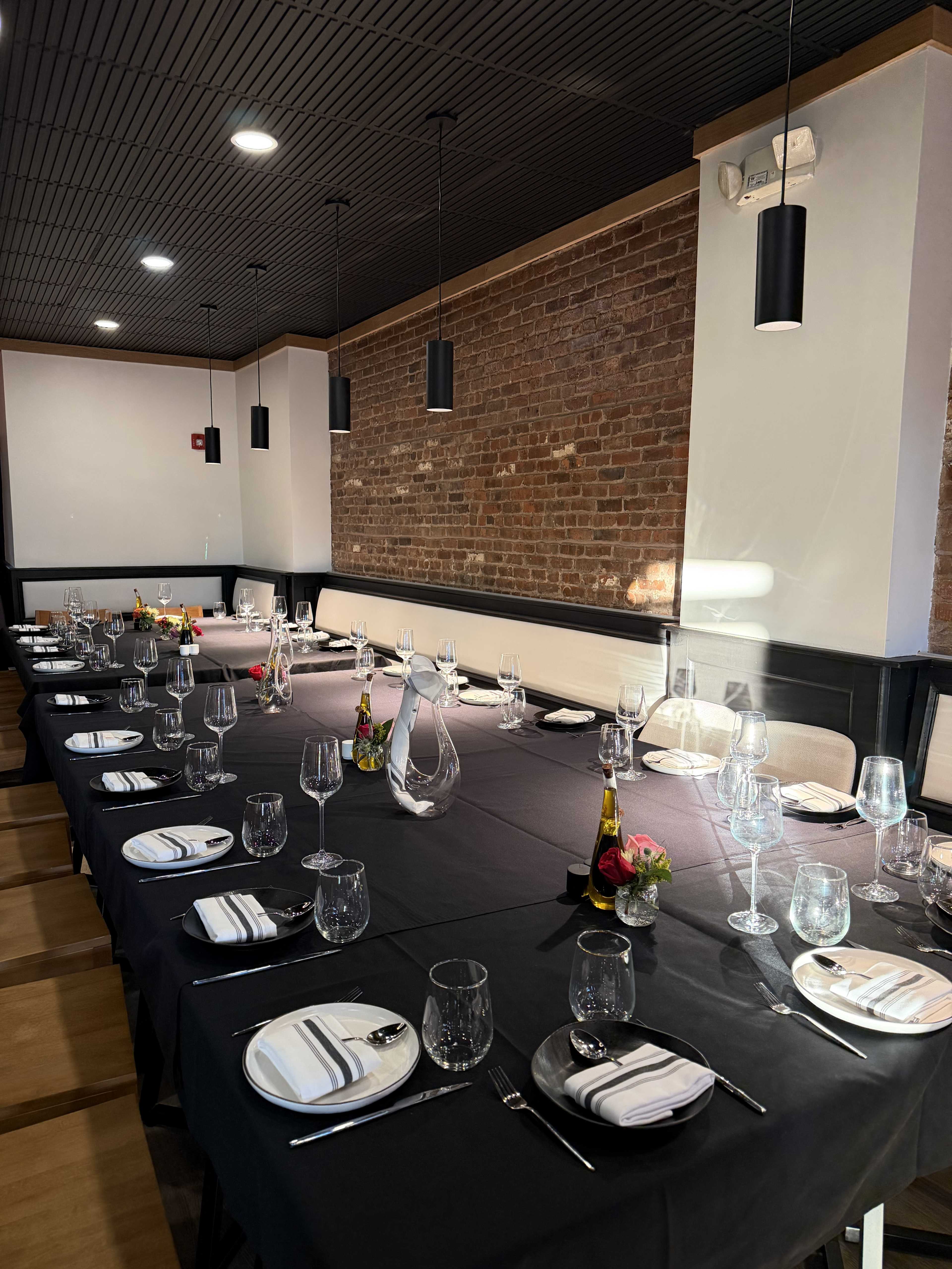 A long dining table is set with glasses, silverware, and floral centerpieces, surrounded by a modern restaurant atmosphere featuring brick walls and pendant lighting.