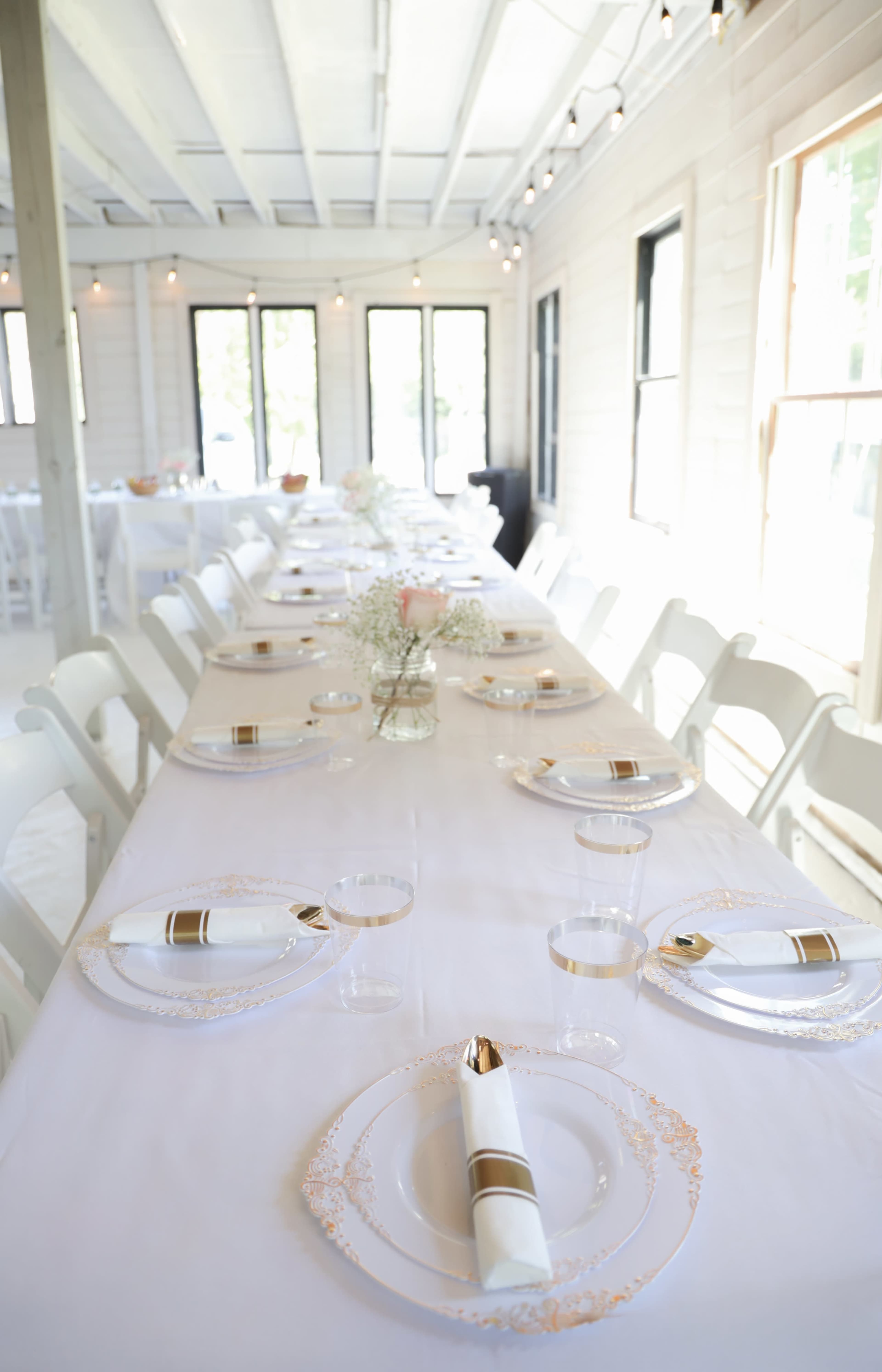 A long dining table is elegantly set with white tablecloths, gold-accented plates, and decorative glassware, in a bright, well-lit indoor space.