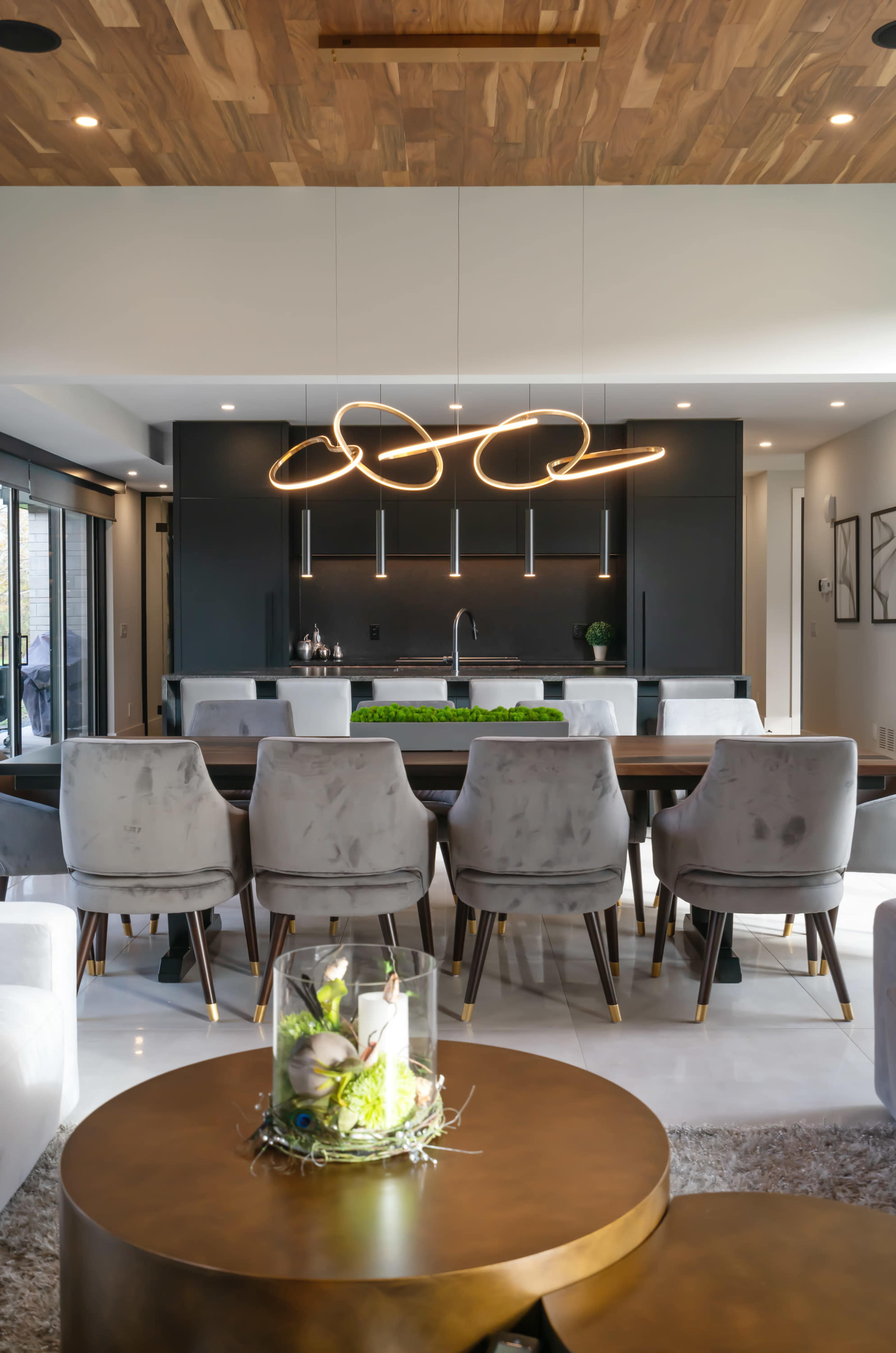 A modern dining area features a long table surrounded by upholstered chairs, with a sleek kitchen in the background and an elegant light fixture overhead.