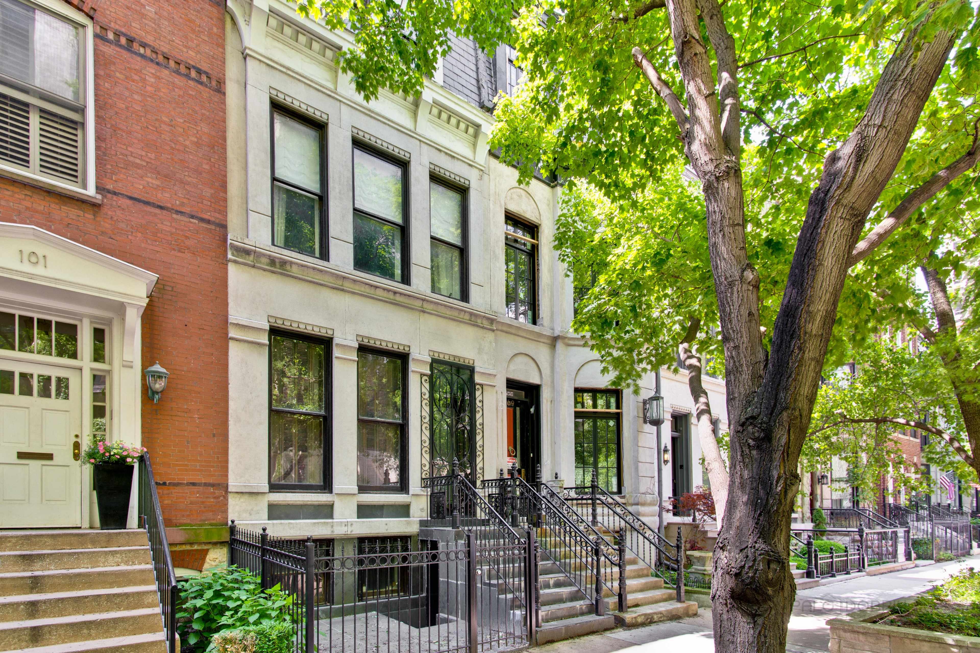 A row of brownstone buildings flanked by lush green trees along a residential sidewalk.