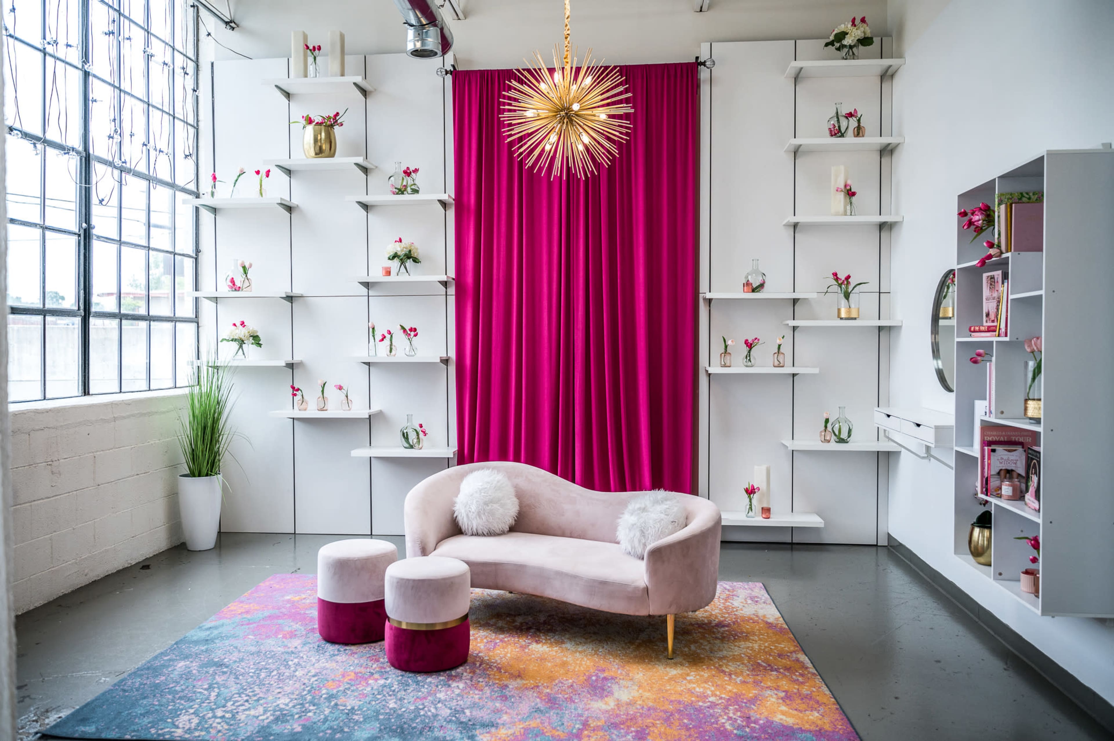 A stylish interior features a pink velvet sofa and matching stools on a colorful rug, set against a backdrop of a bright pink curtain and white shelving displaying decorative flowers.