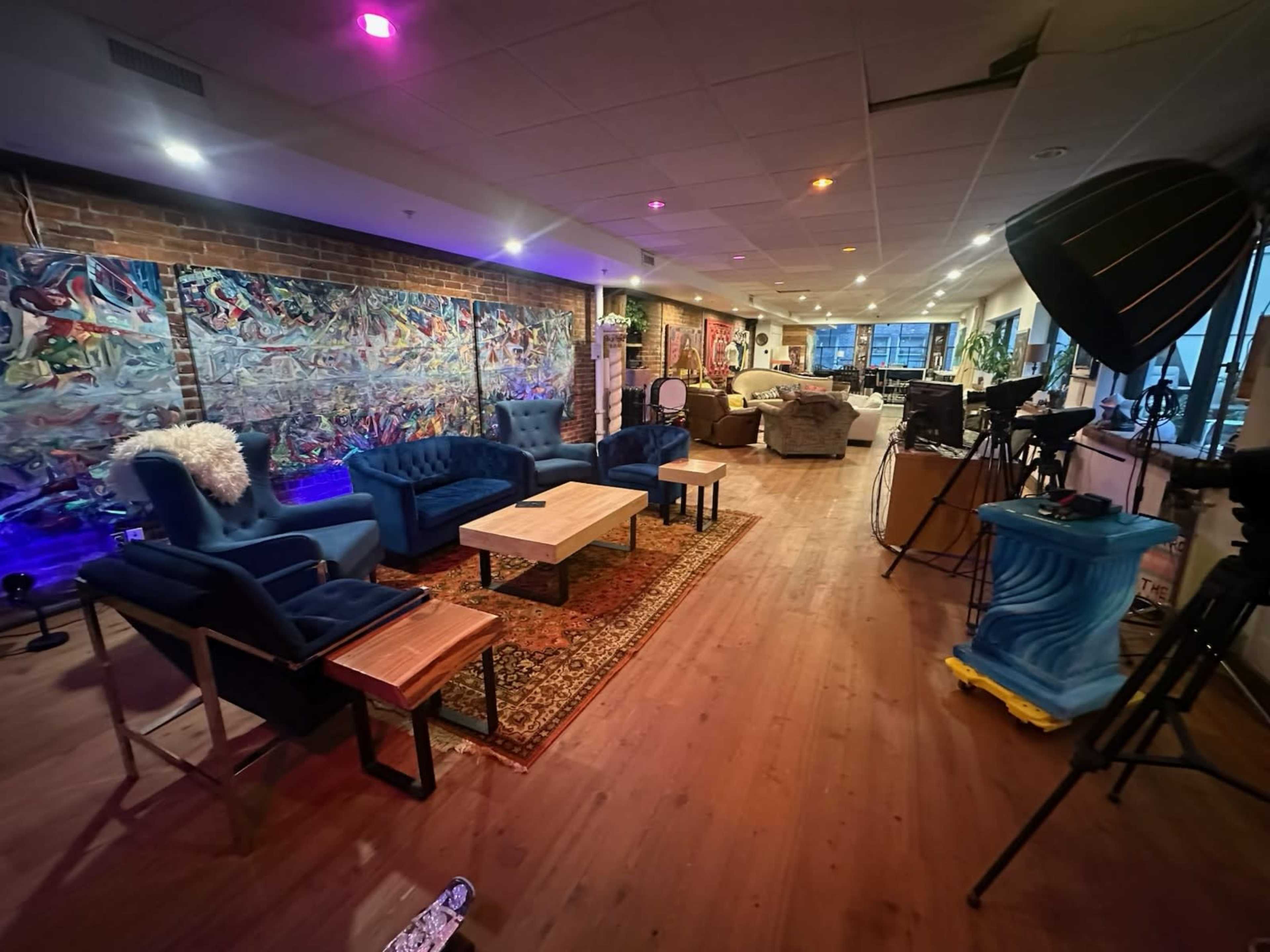 The image depicts a spacious indoor area with multiple seating arrangements, including blue chairs and wooden tables, alongside various decorations and filming equipment.