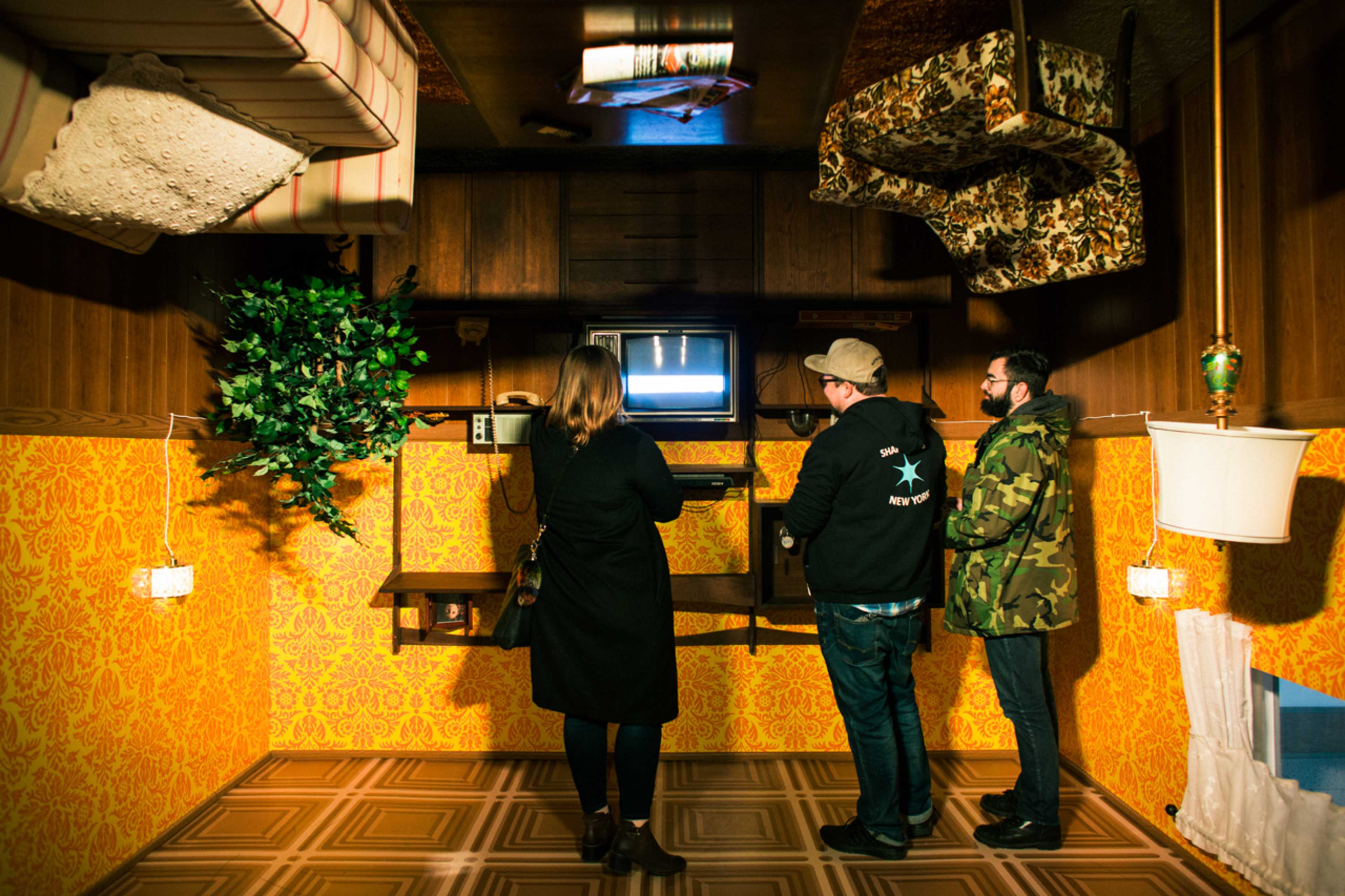 Three people stand in a room with upside-down furniture, watching a television screen.