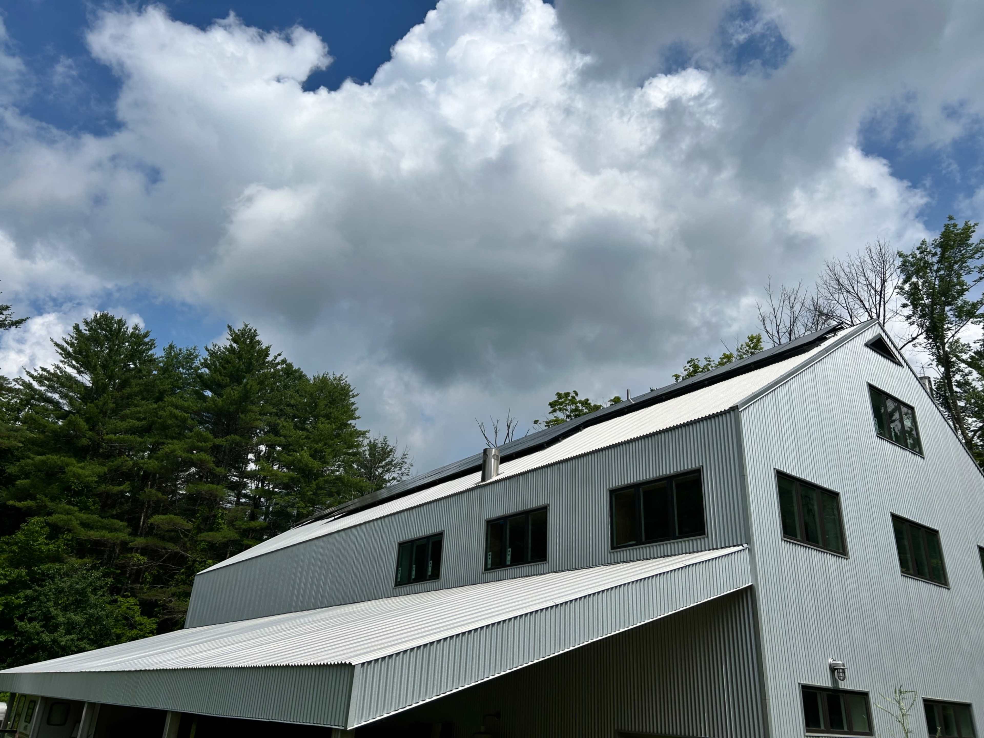 A metal-roofed building with large windows is situated beneath a cloudy sky and surrounded by trees.