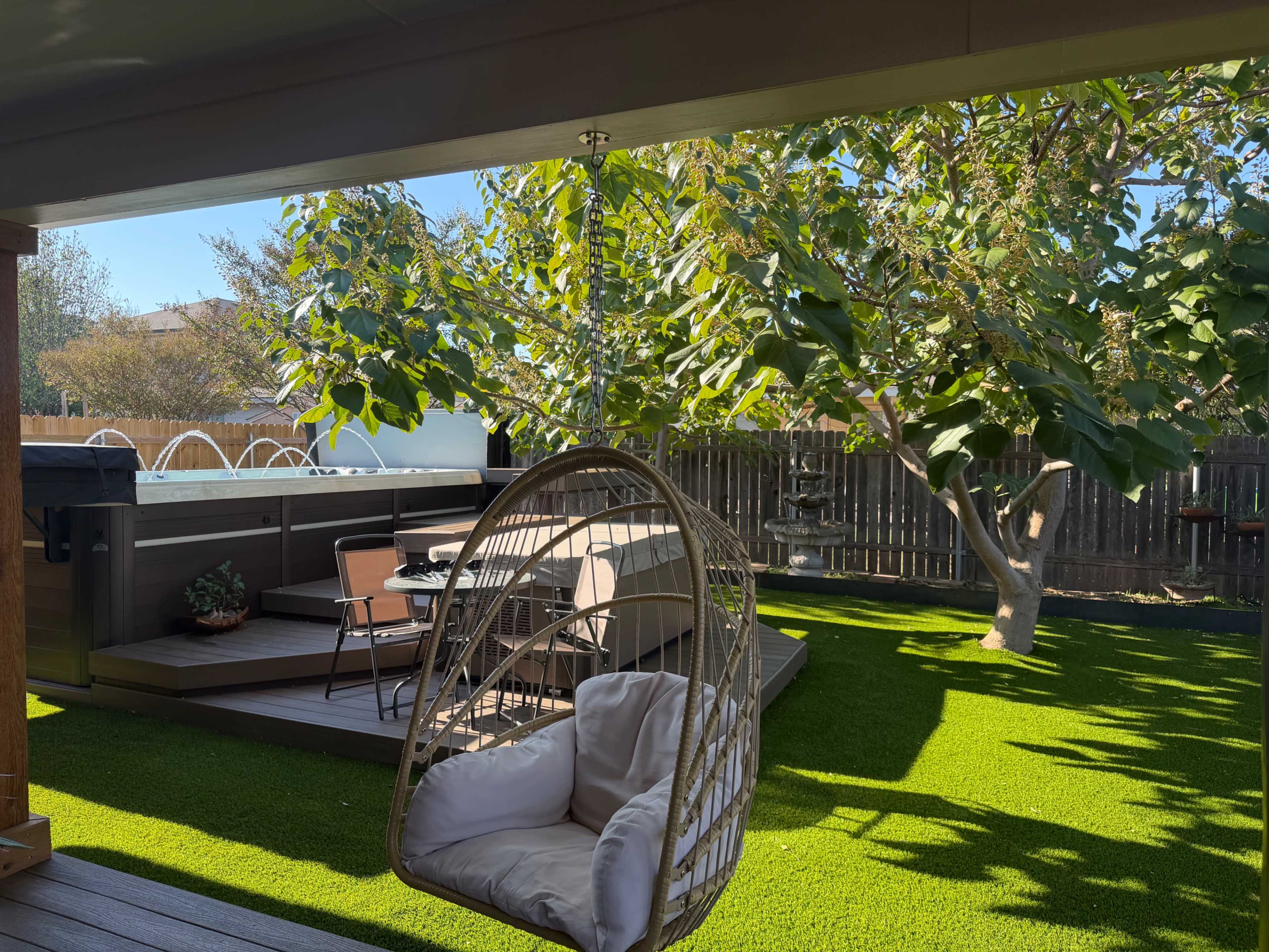 The image shows a backyard with a hanging chair, a patio set, and a hot tub surrounded by greenery and a wooden fence.