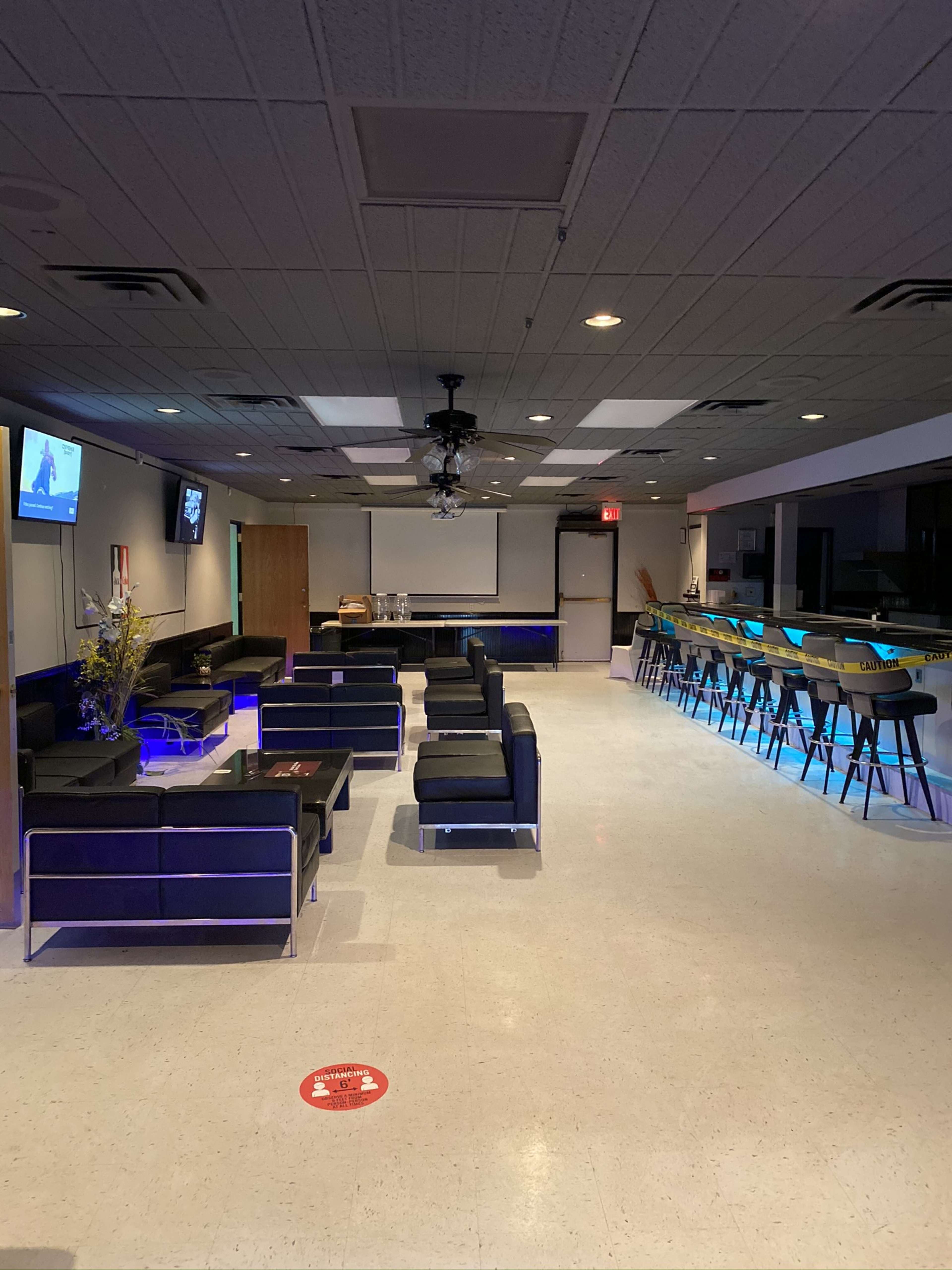 The image shows a spacious interior of a lounge area featuring a seating arrangement with dark couches and a bar with illuminated stools.