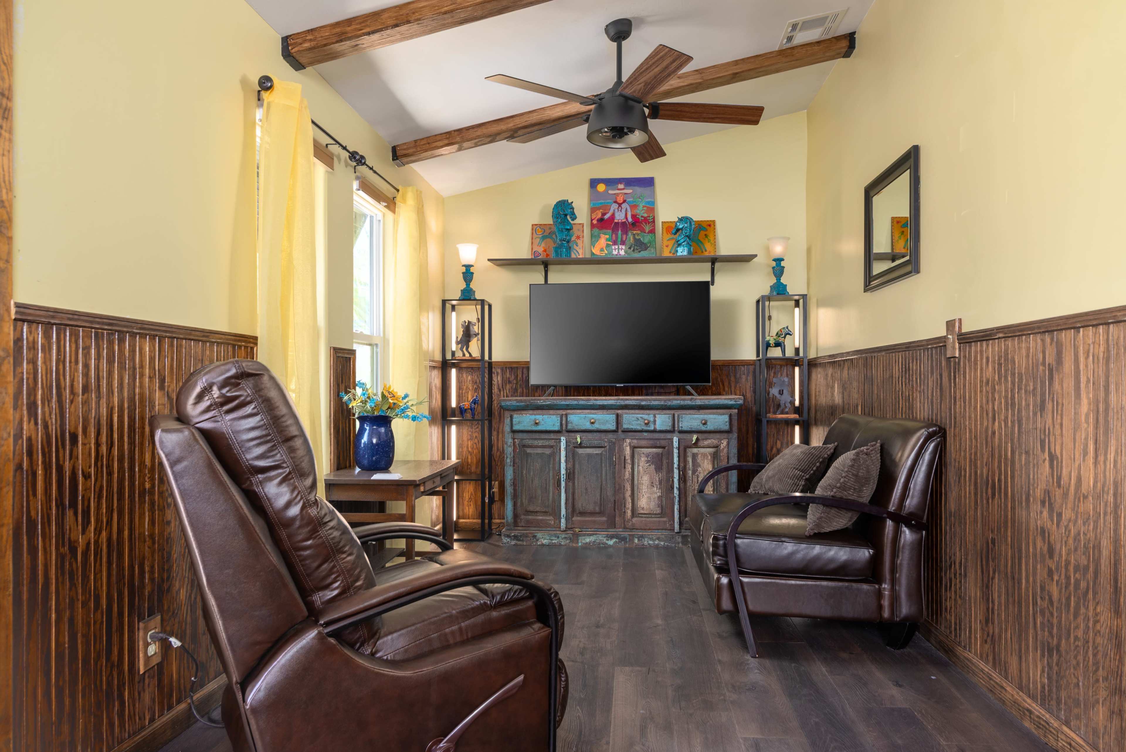 The image shows a living room with wooden beams on the ceiling, a television mounted on the wall, and two leather chairs situated in front of a rustic wooden cabinet.
