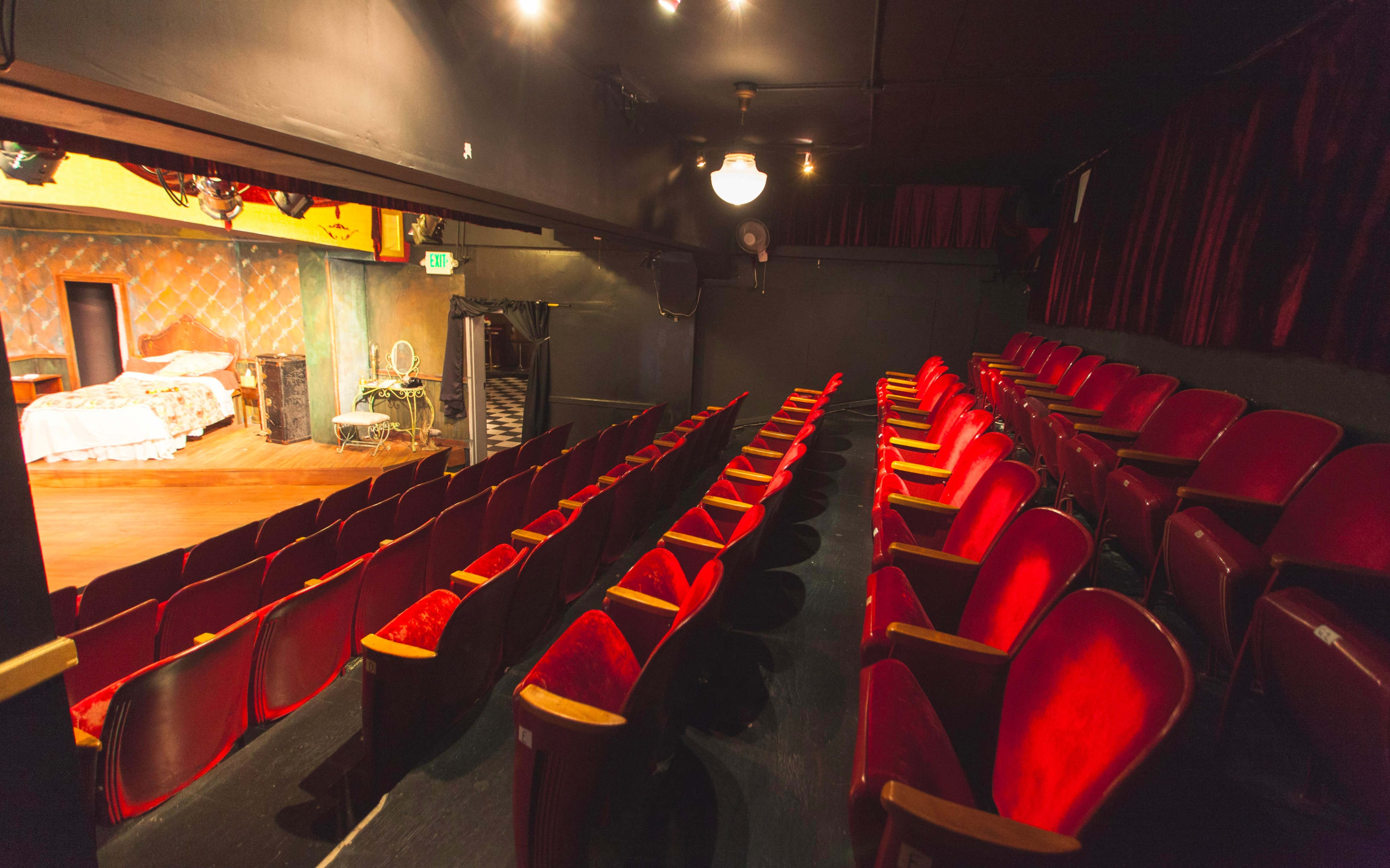 An empty theater with red seats facing a small stage that features a bed and a vanity.