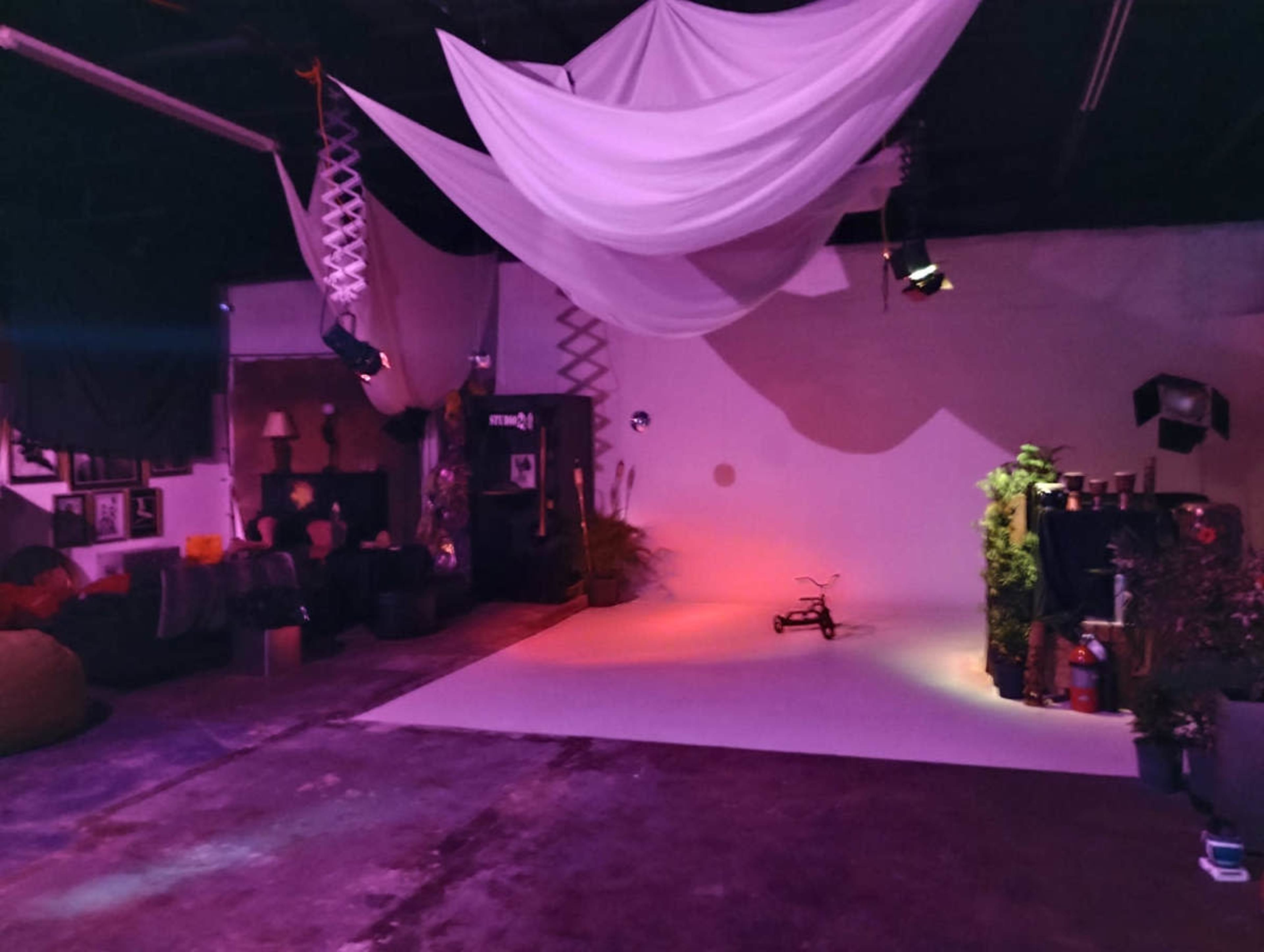 The image shows a spacious, dimly lit studio with draped fabric, various lighting fixtures, and a minimalist backdrop set for a photo shoot.