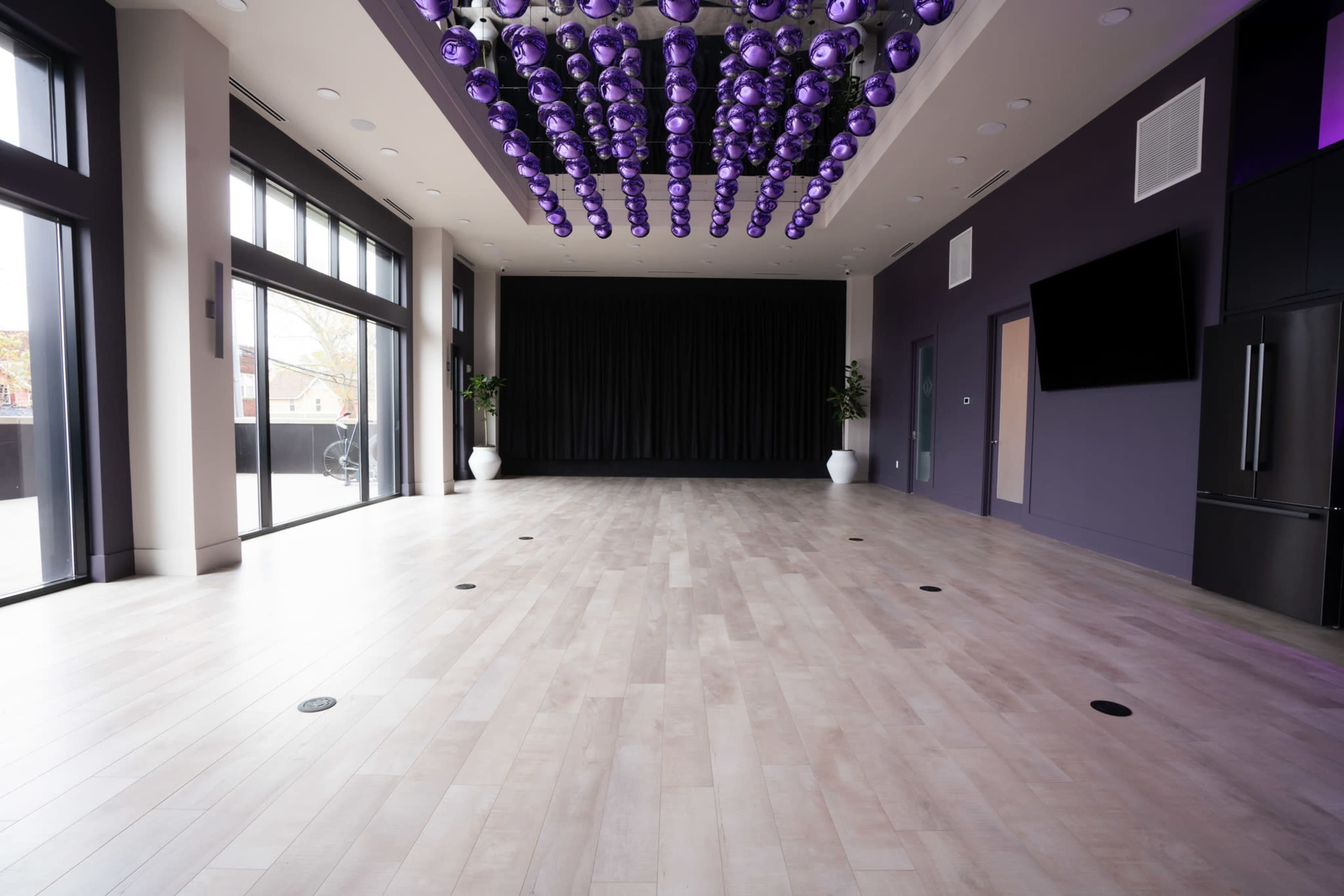 A spacious room with light wood flooring, large windows, and a decorative installation of purple spheres hanging from the ceiling.