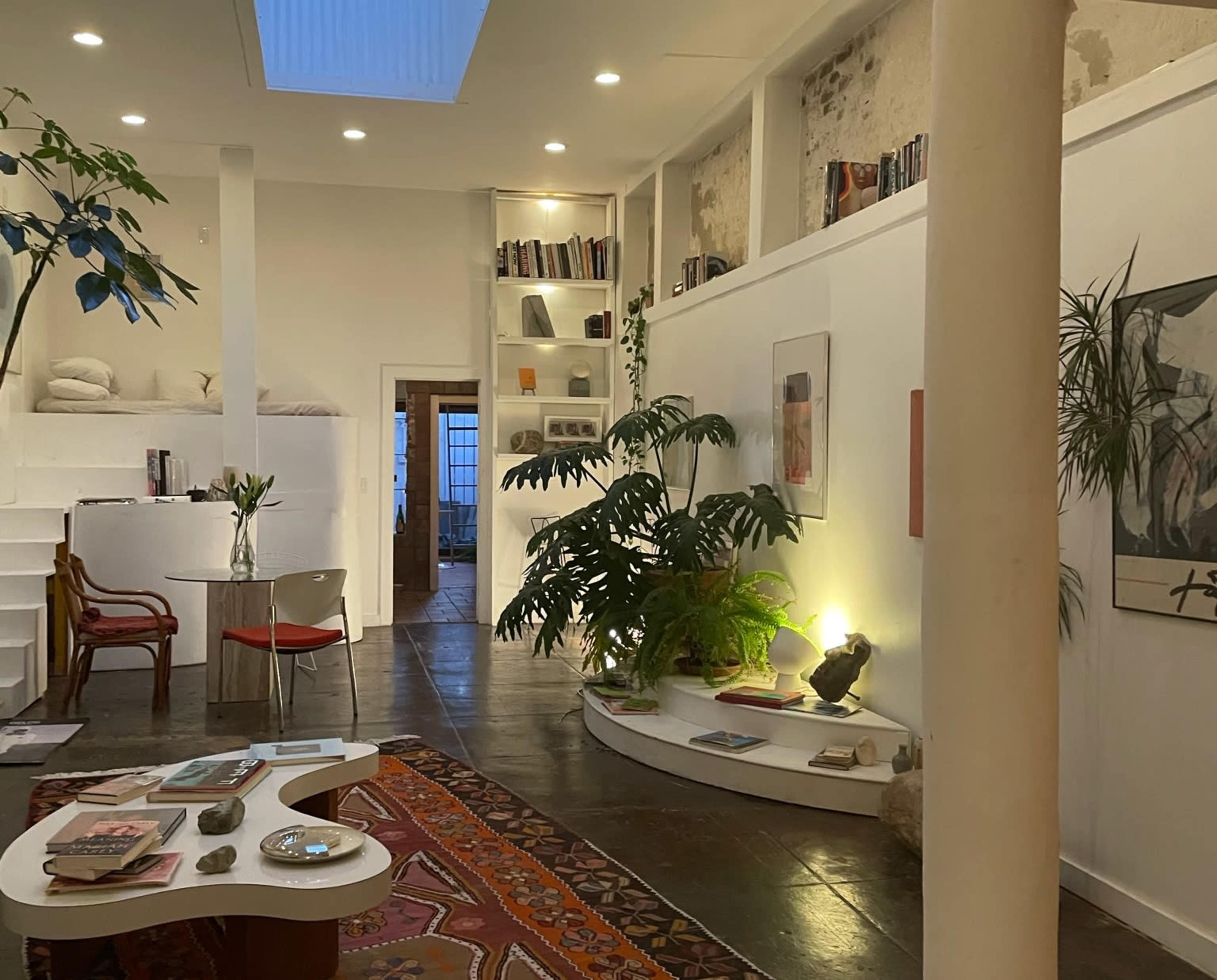 The image shows a cozy interior space with a spiral staircase, bookshelves, plants, and a circular coffee table on a patterned rug.