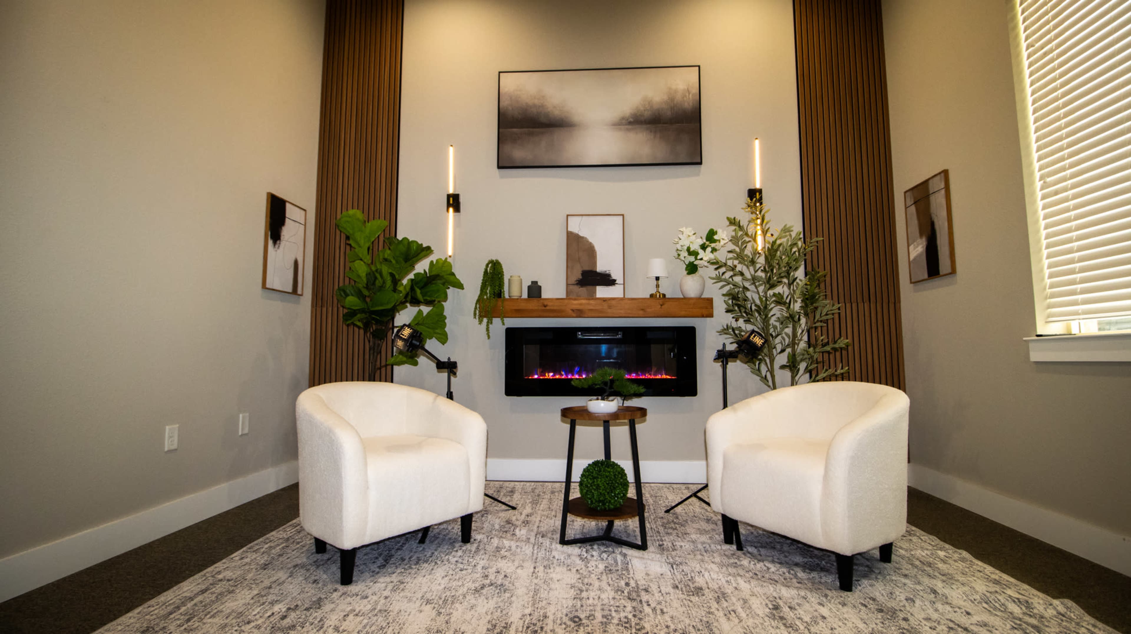 The image features a cozy seating area with two white chairs and a modern electric fireplace surrounded by decorative plants and wall art.