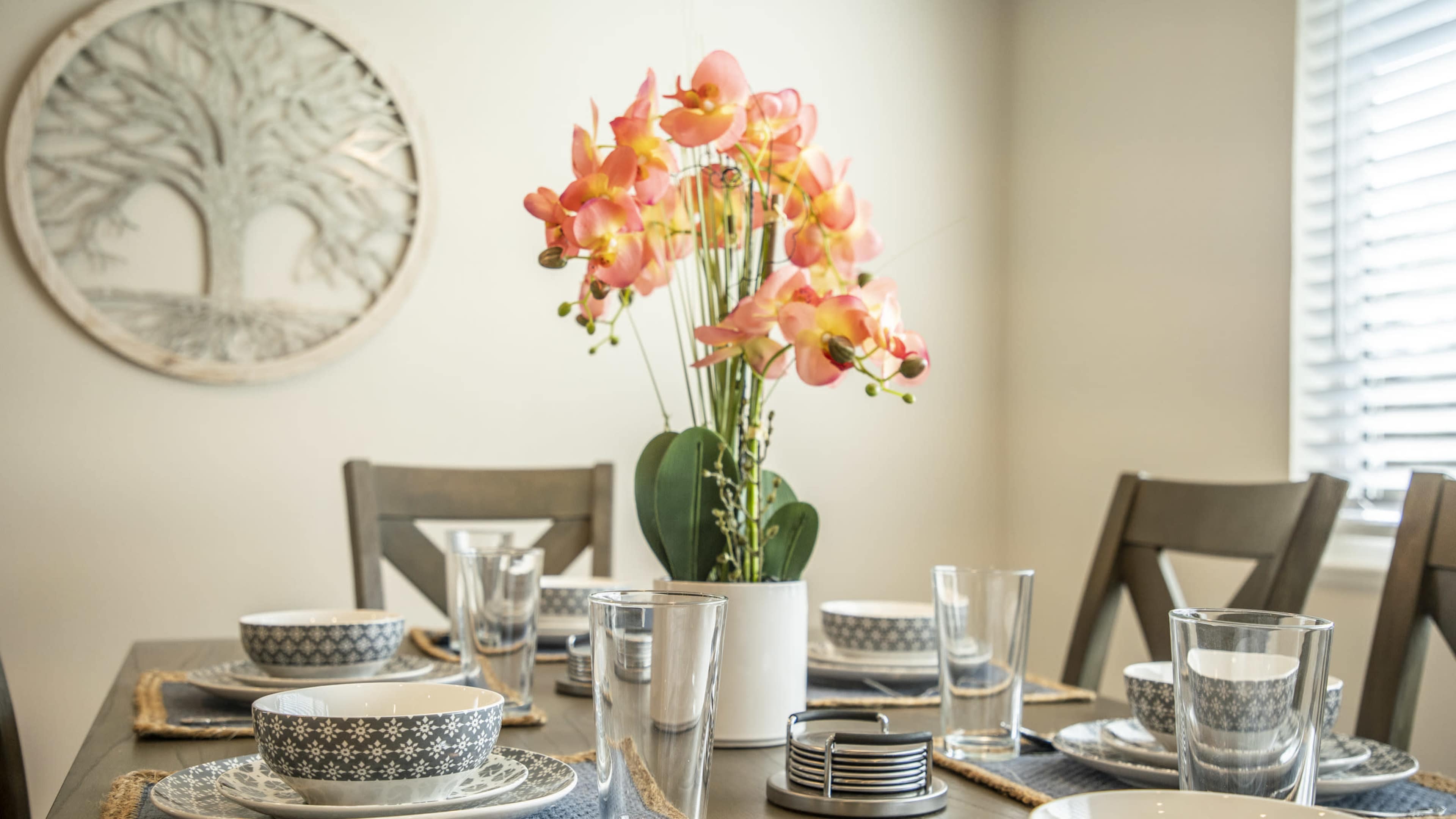 A dining table is set with plates, glasses, and a centerpiece of orchids, accompanied by a decorative wall piece featuring trees.