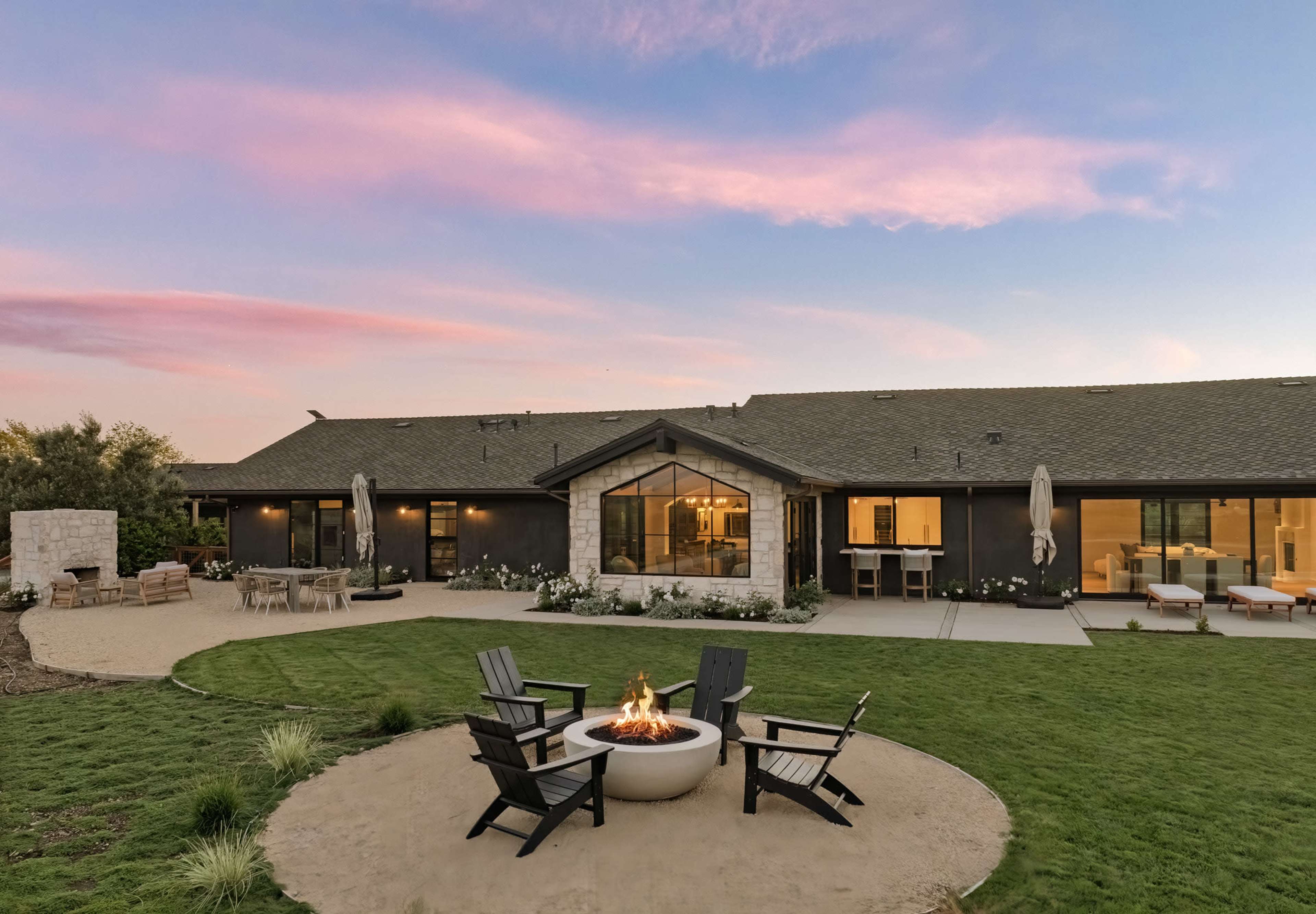 A backyard features a circular fire pit surrounded by four chairs, with a house in the background under a colorful sunset sky.