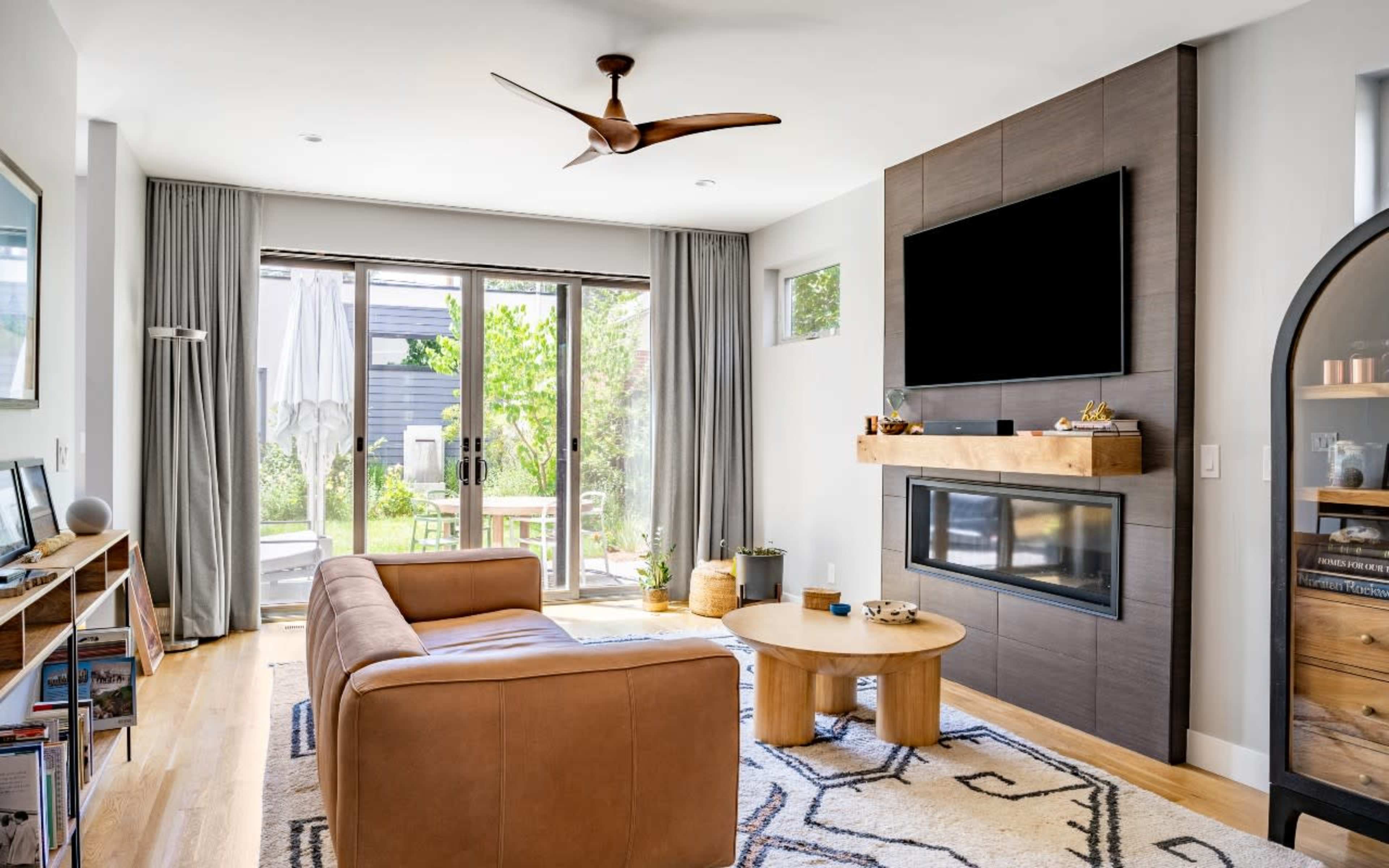 A modern living room features a tan leather sofa, a round wooden coffee table, and a wall-mounted television above a fireplace, with large windows opening to a landscaped outdoor area.