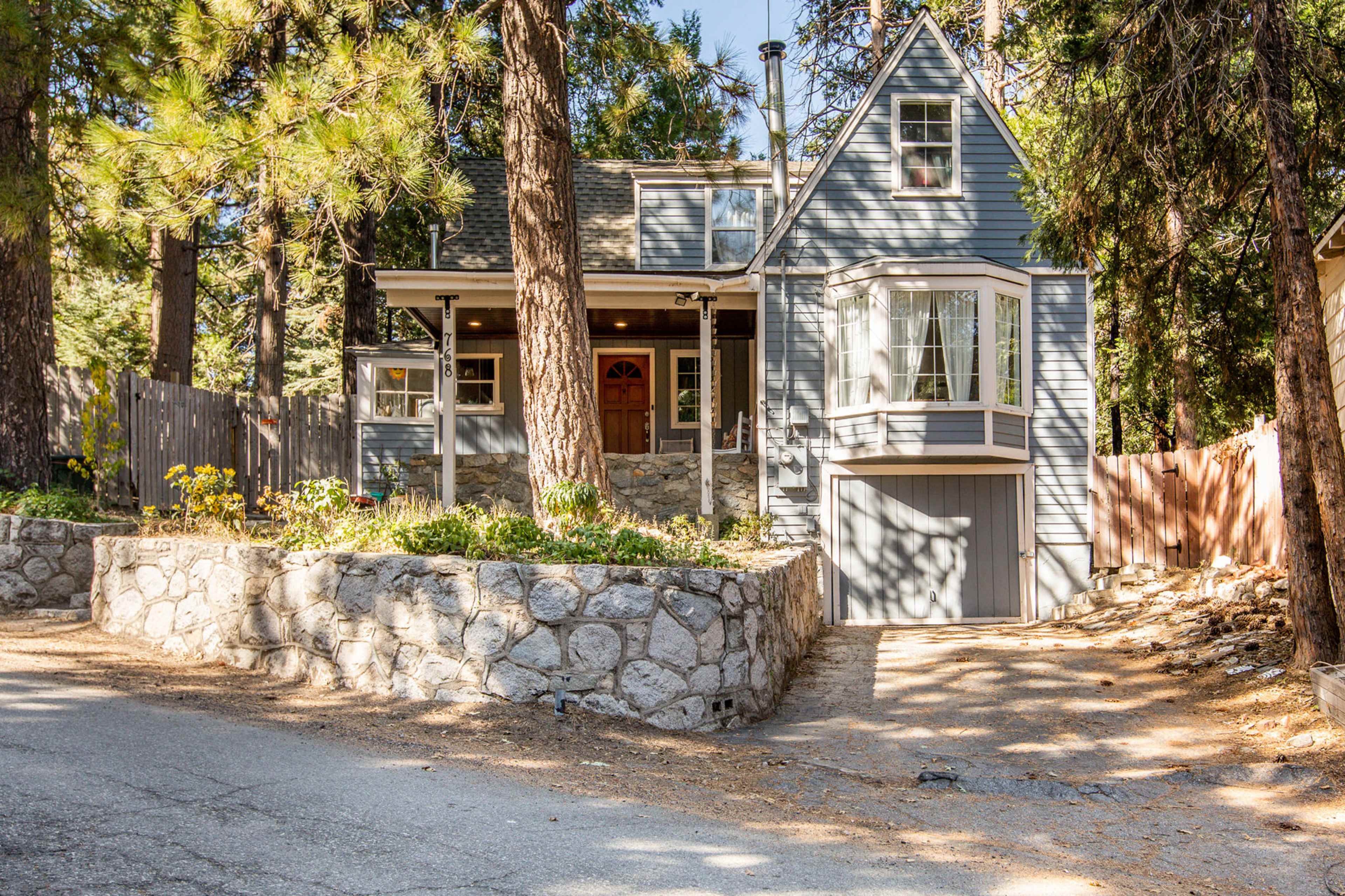 A gray two-story house with a stone foundation is situated on a tree-lined street.