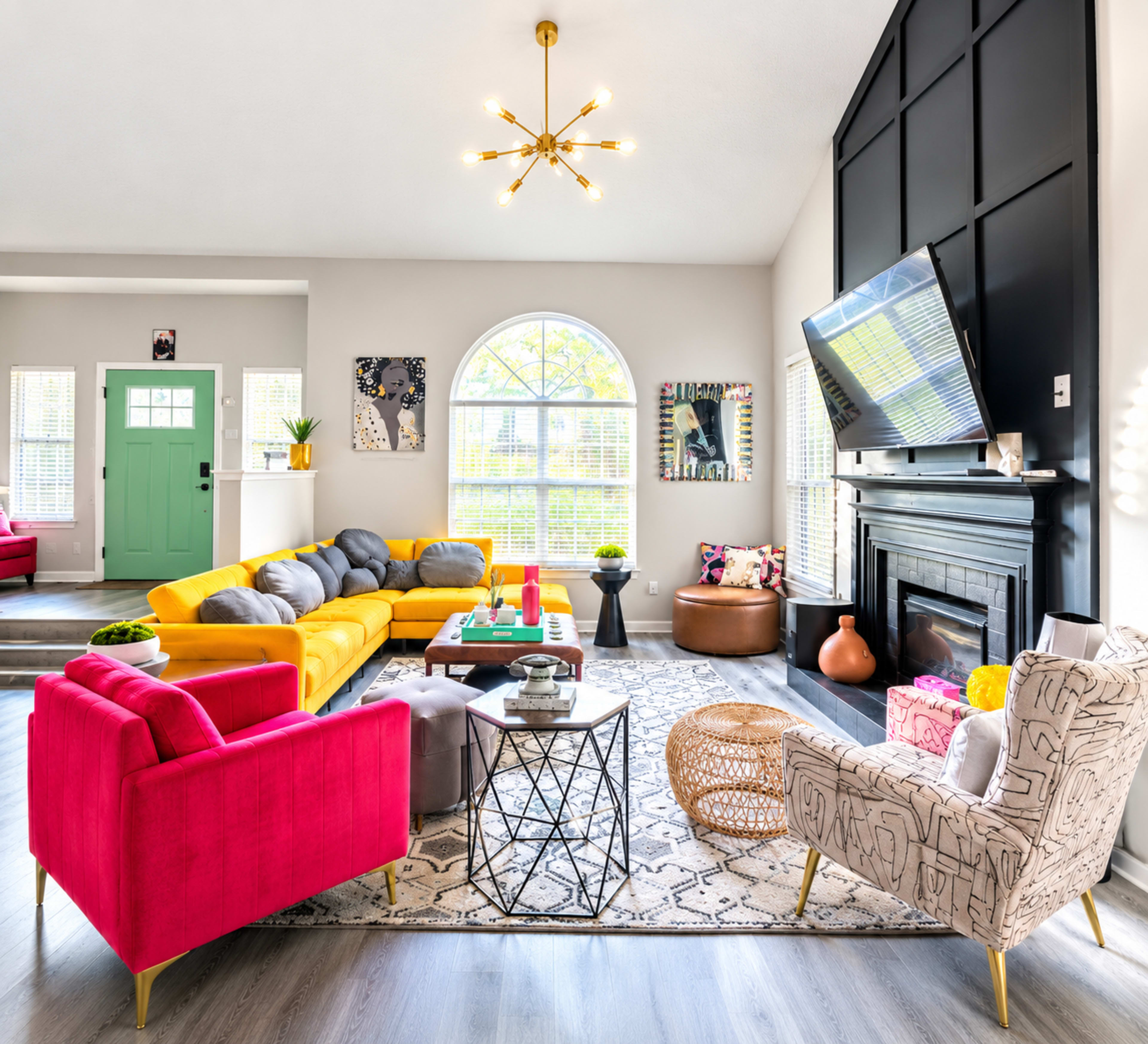 The image shows a bright and modern living room featuring a yellow sectional sofa, a pink armchair, a patterned rug, and various decorative elements.