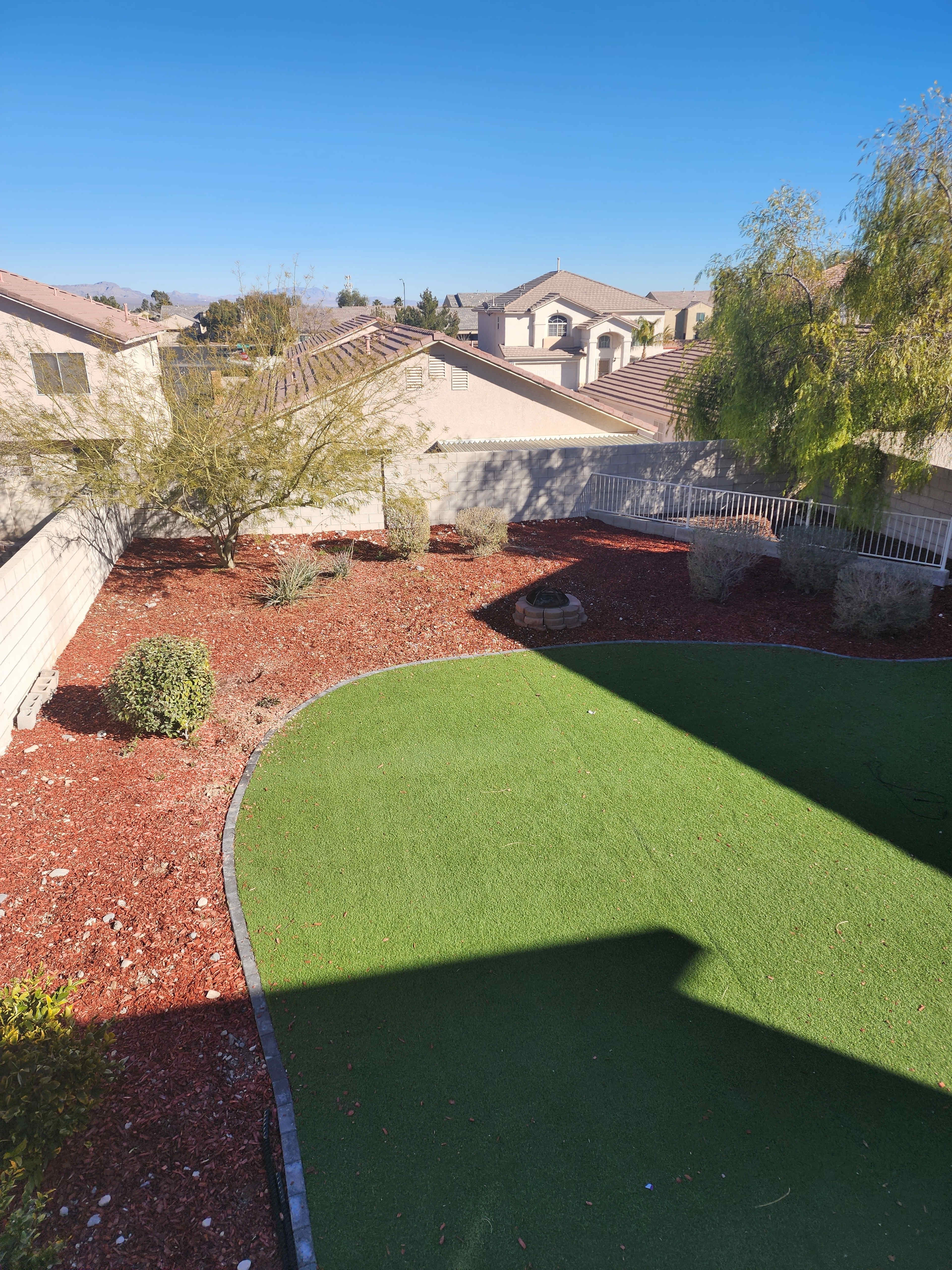A backyard with artificial grass, landscaped areas with shrubs, and a view of neighboring homes in the background.