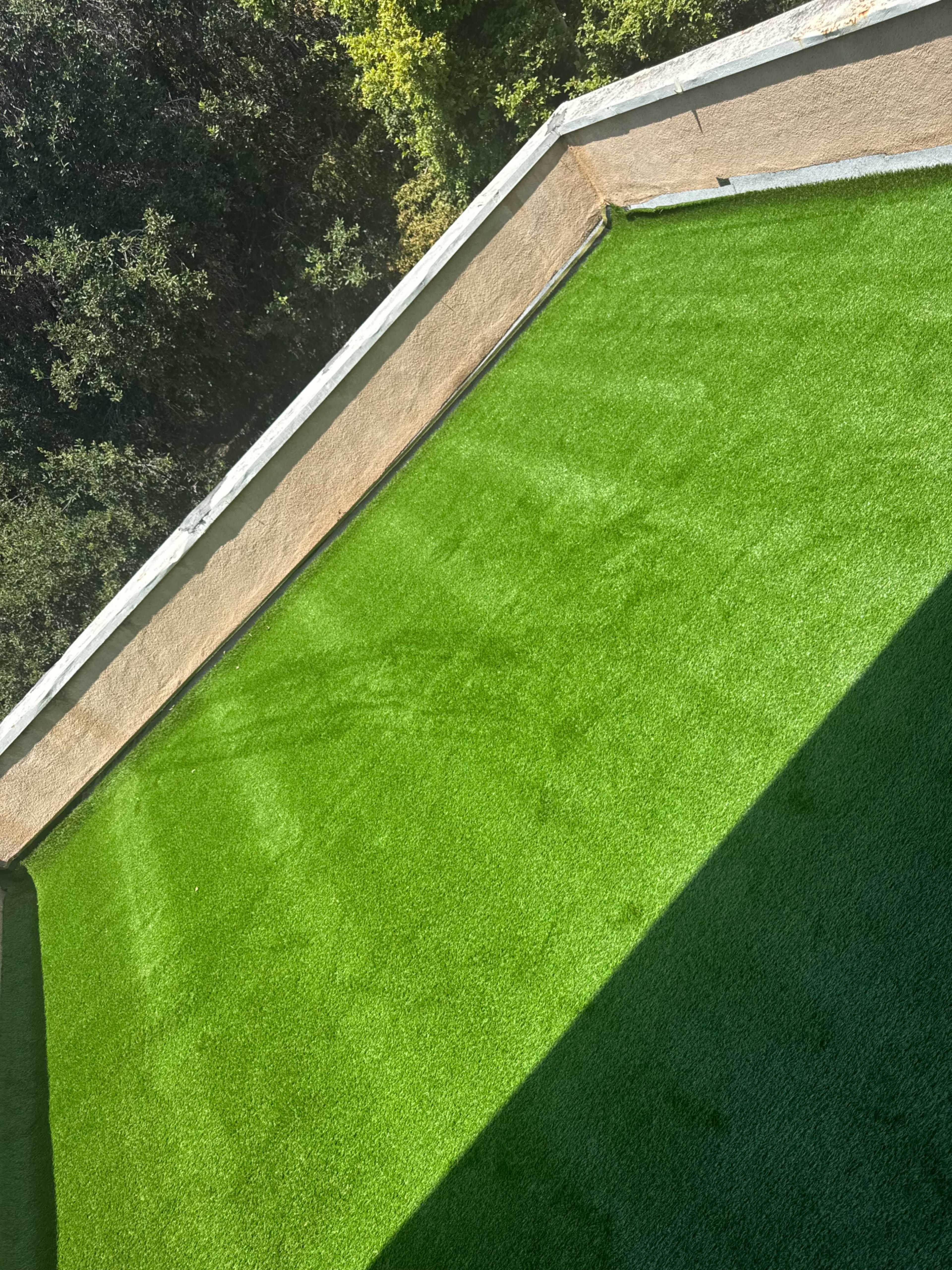 The image shows a flat area covered with bright green artificial grass, bordered by a tan wall and surrounded by trees in the background.