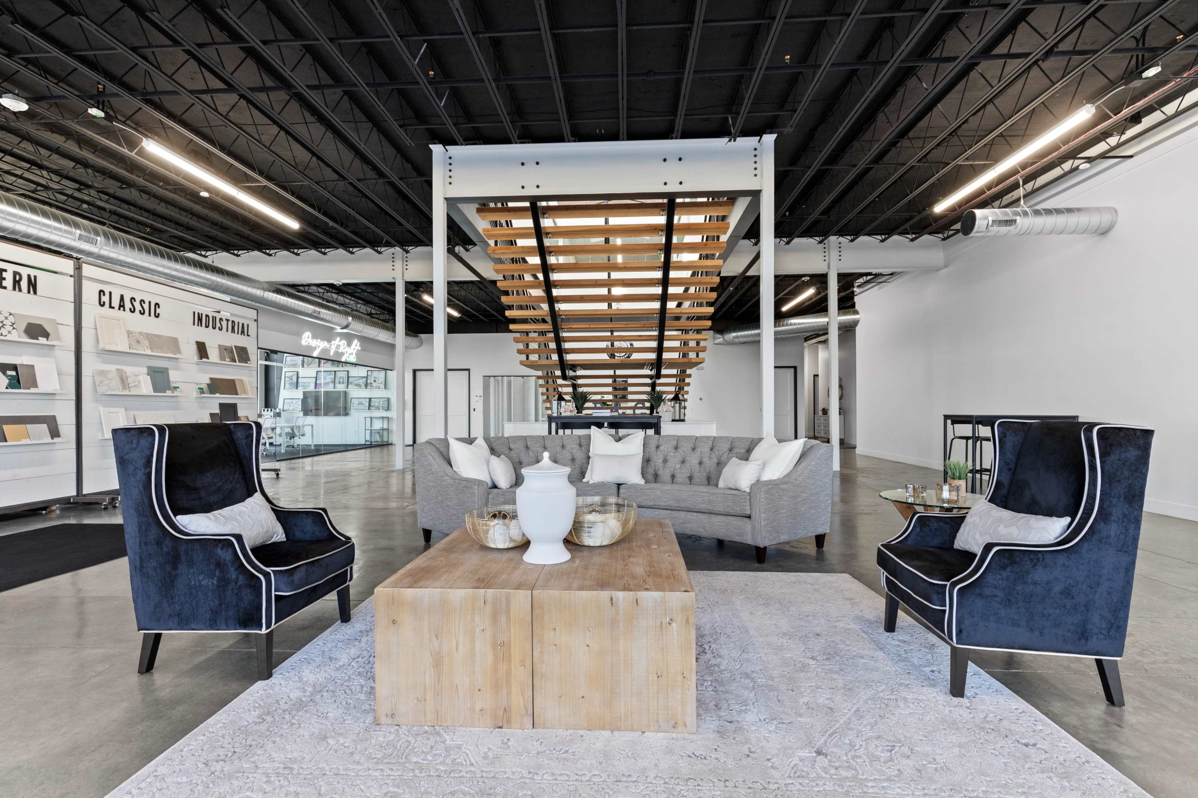 The image shows a modern interior space featuring a seating area with a gray sofa, two blue armchairs, and a wooden coffee table under a staircase.