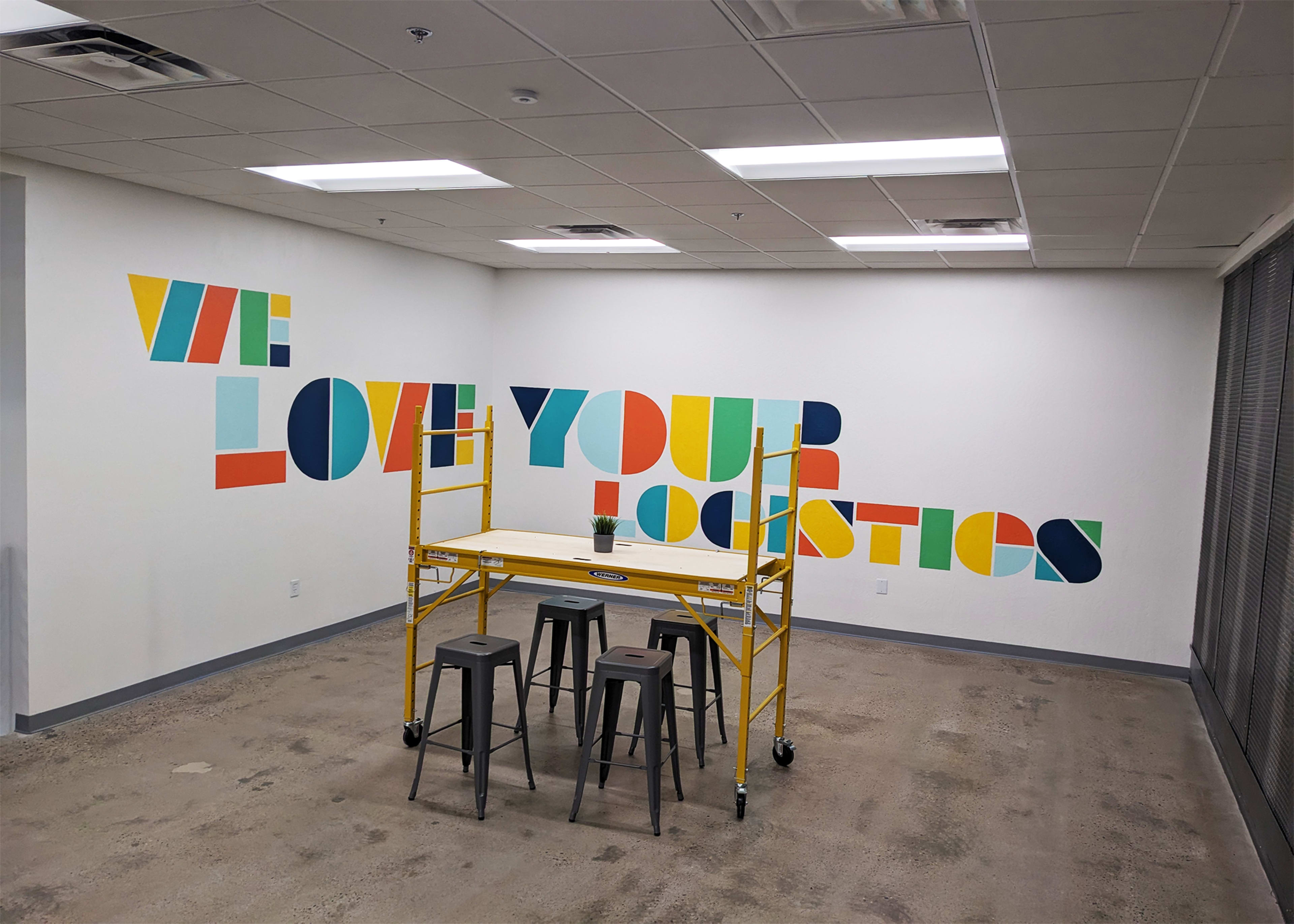 An empty office space with a yellow scaffold and three black stools in front of a colorful wall mural that reads "WE LOVE YOUR LOGISTICS."