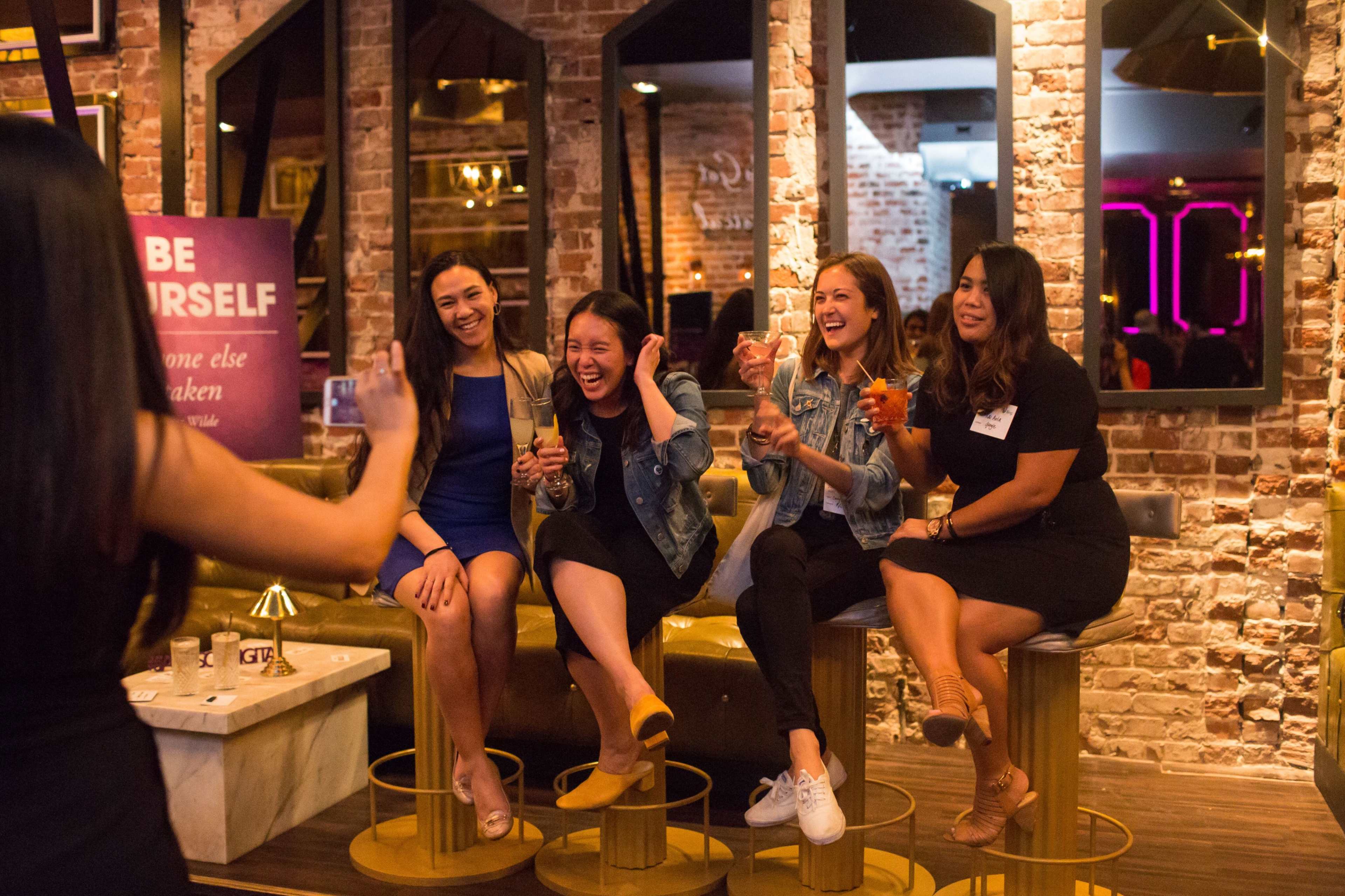 Four women are sitting on bar stools, smiling and laughing while posing for a photo in a lively bar setting with exposed brick walls.