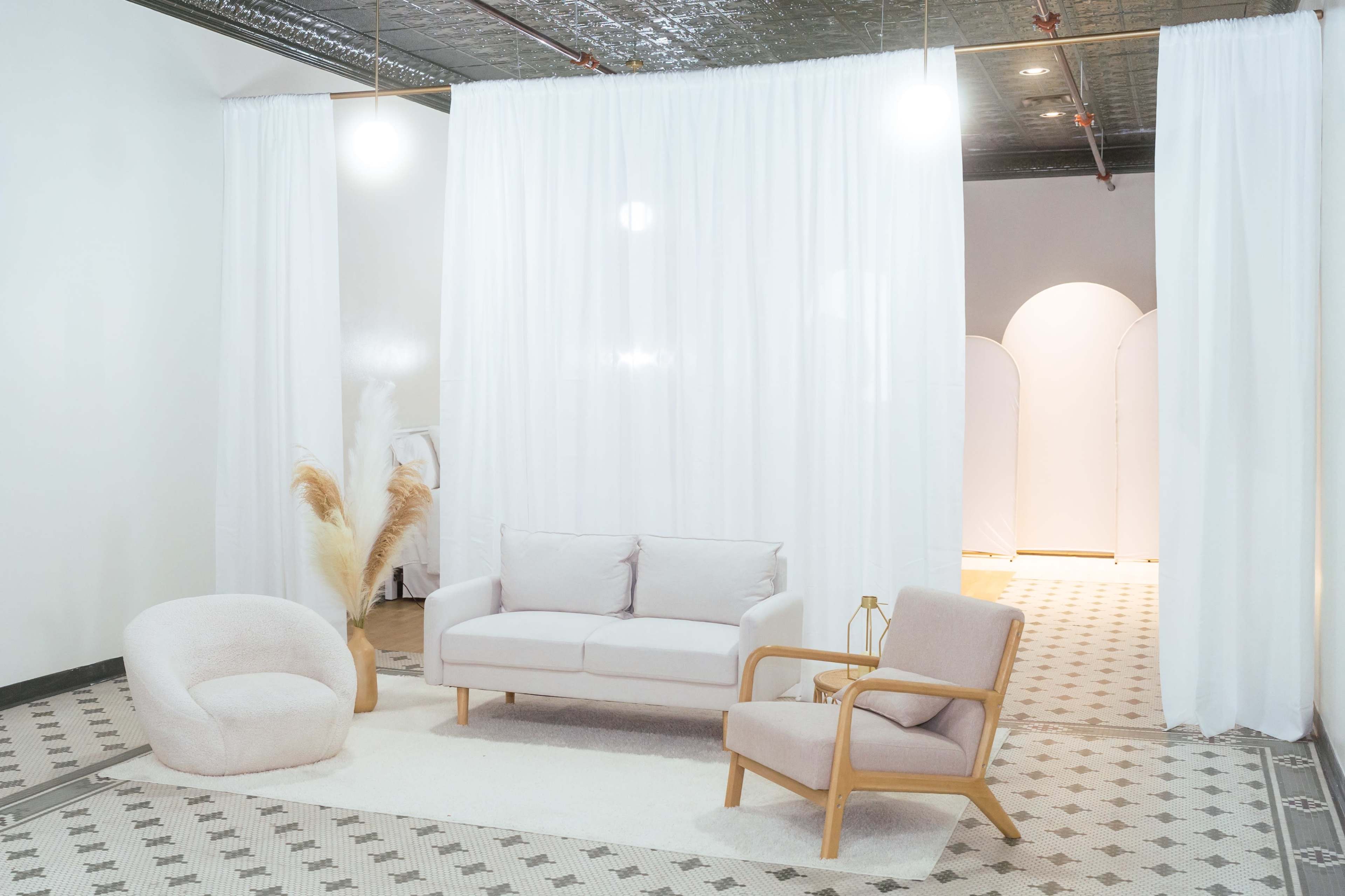 The image depicts a minimalistic interior space with a light-colored sofa, armchair, and a round chair, separated by white curtains in a room with patterned flooring.
