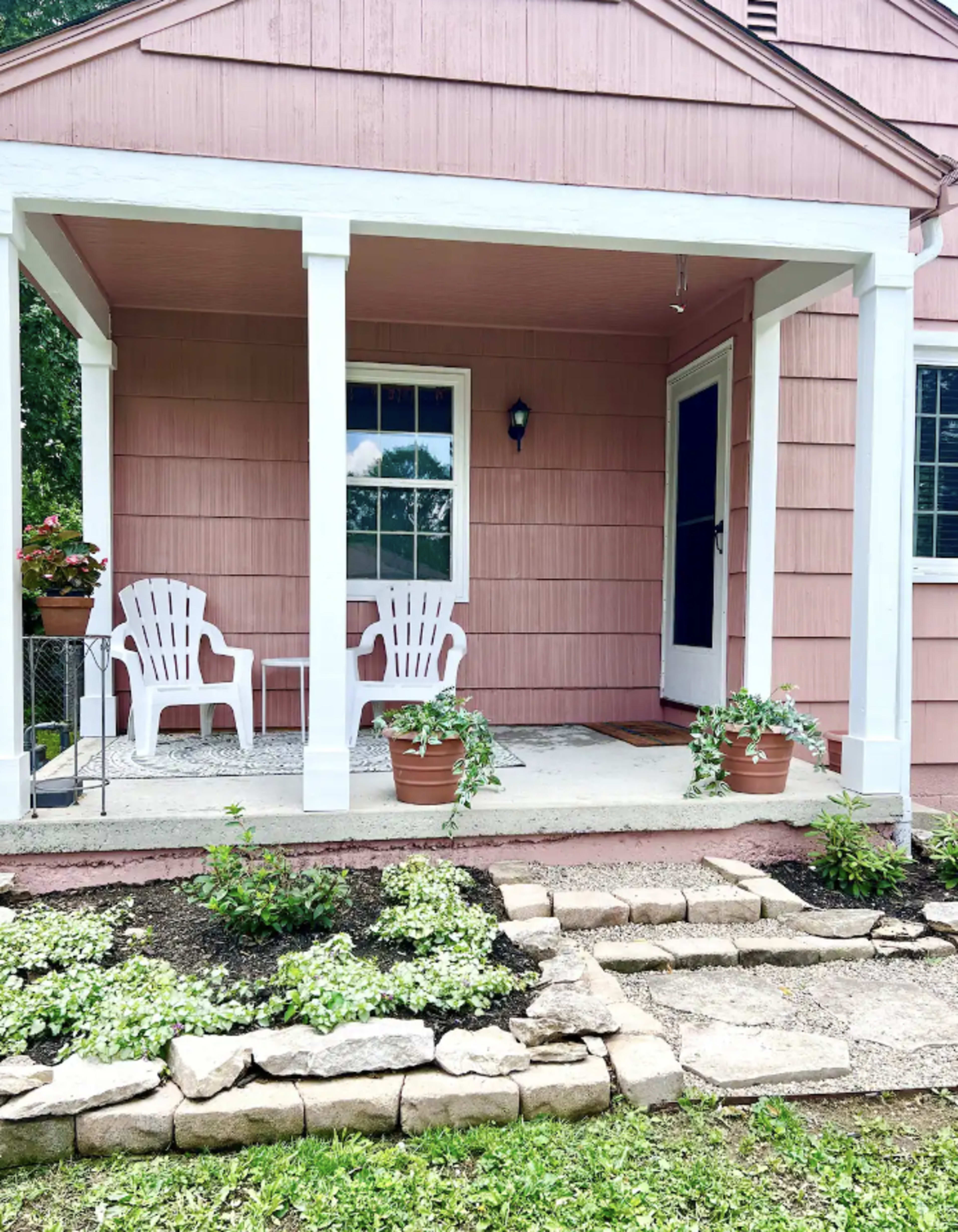 A pink house with a front porch featuring two white chairs, a potted plant, and a pathway made of stones leading to the entrance.