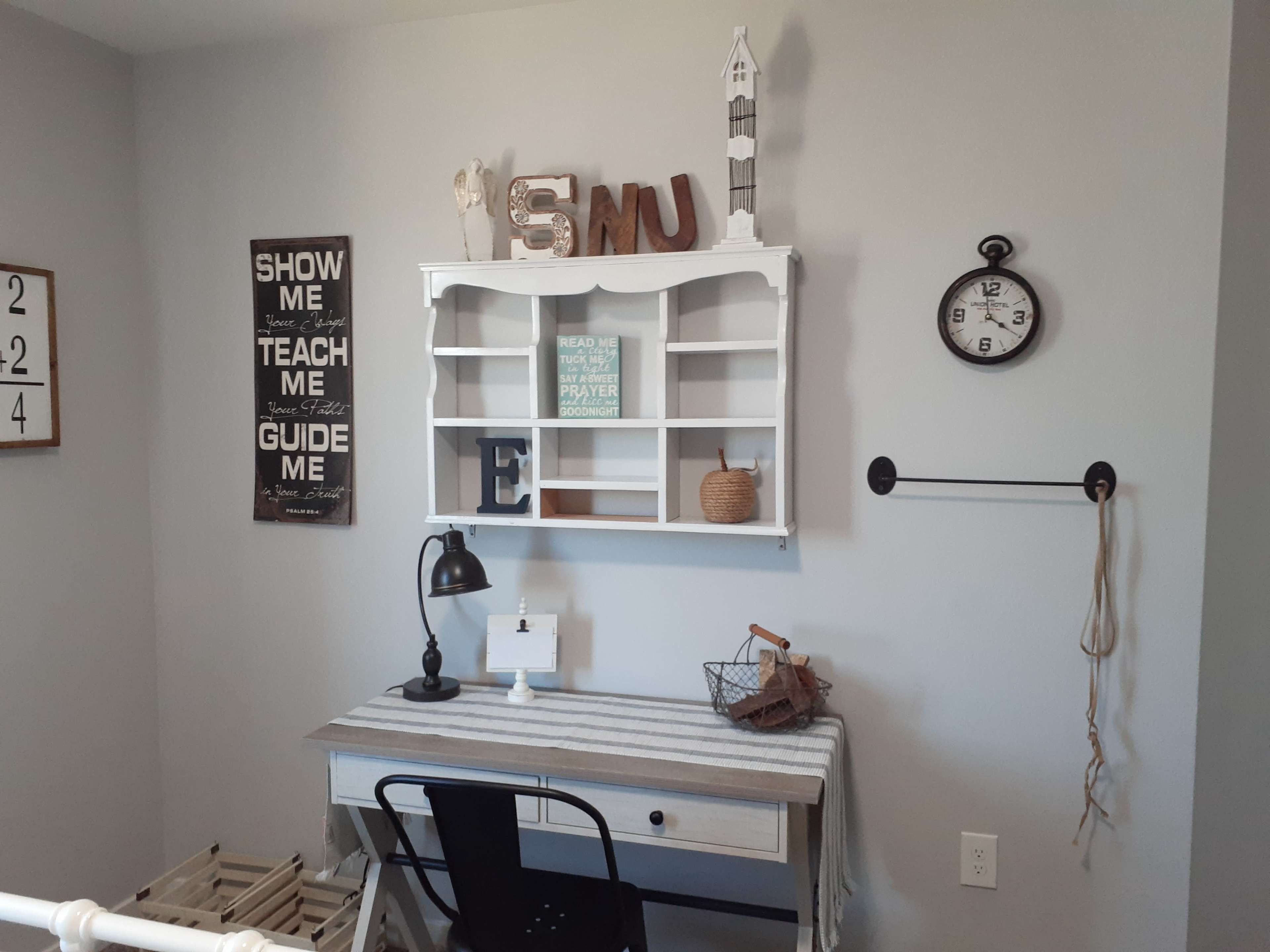 A small, organized workspace with a white desk, black chair, and decorative wall shelves displaying various items and a clock.