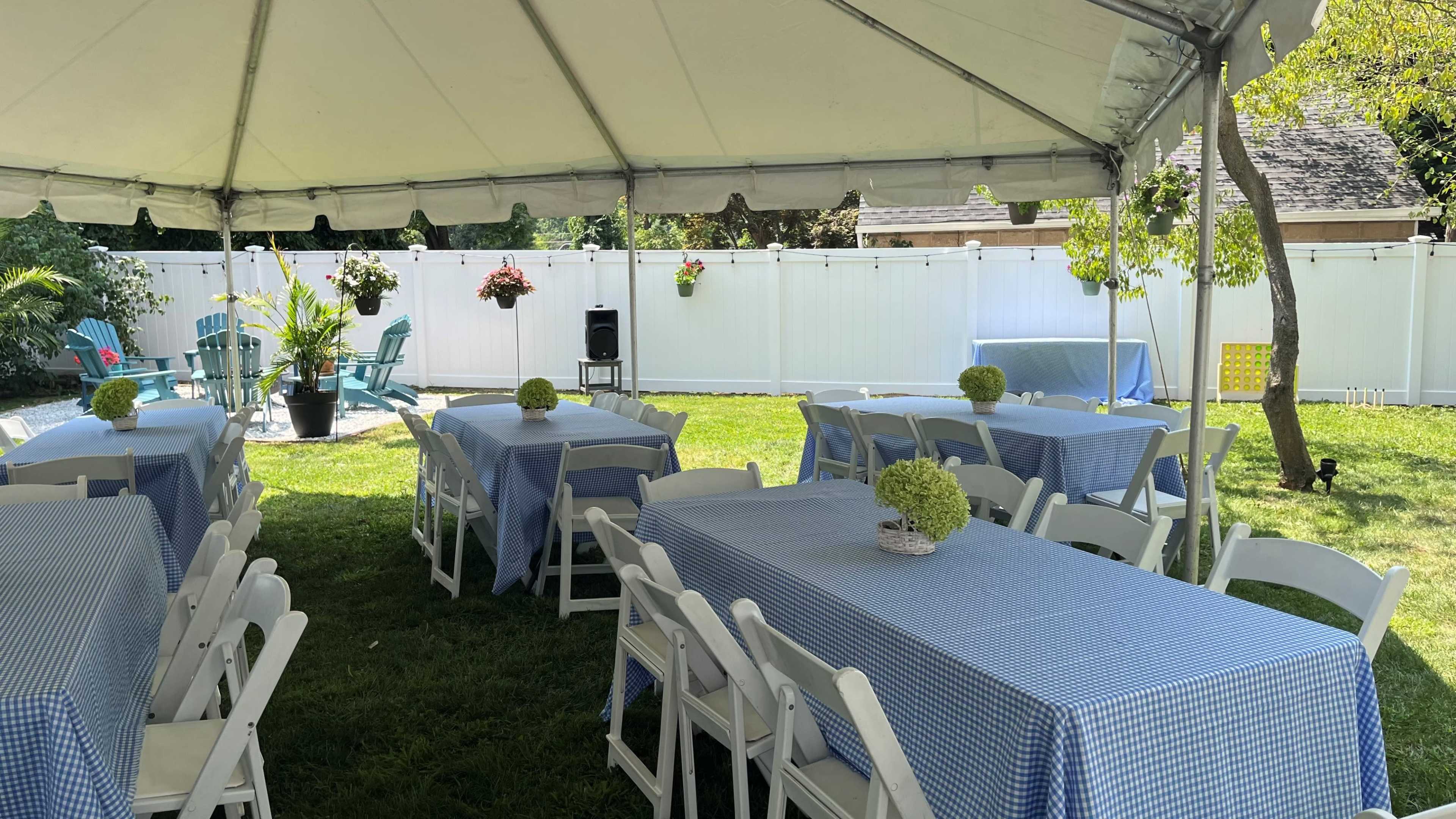A large tent with tables covered in blue checkered tablecloths is set up on a grassy area, surrounded by white fencing and decorated with potted plants and hanging flowers.