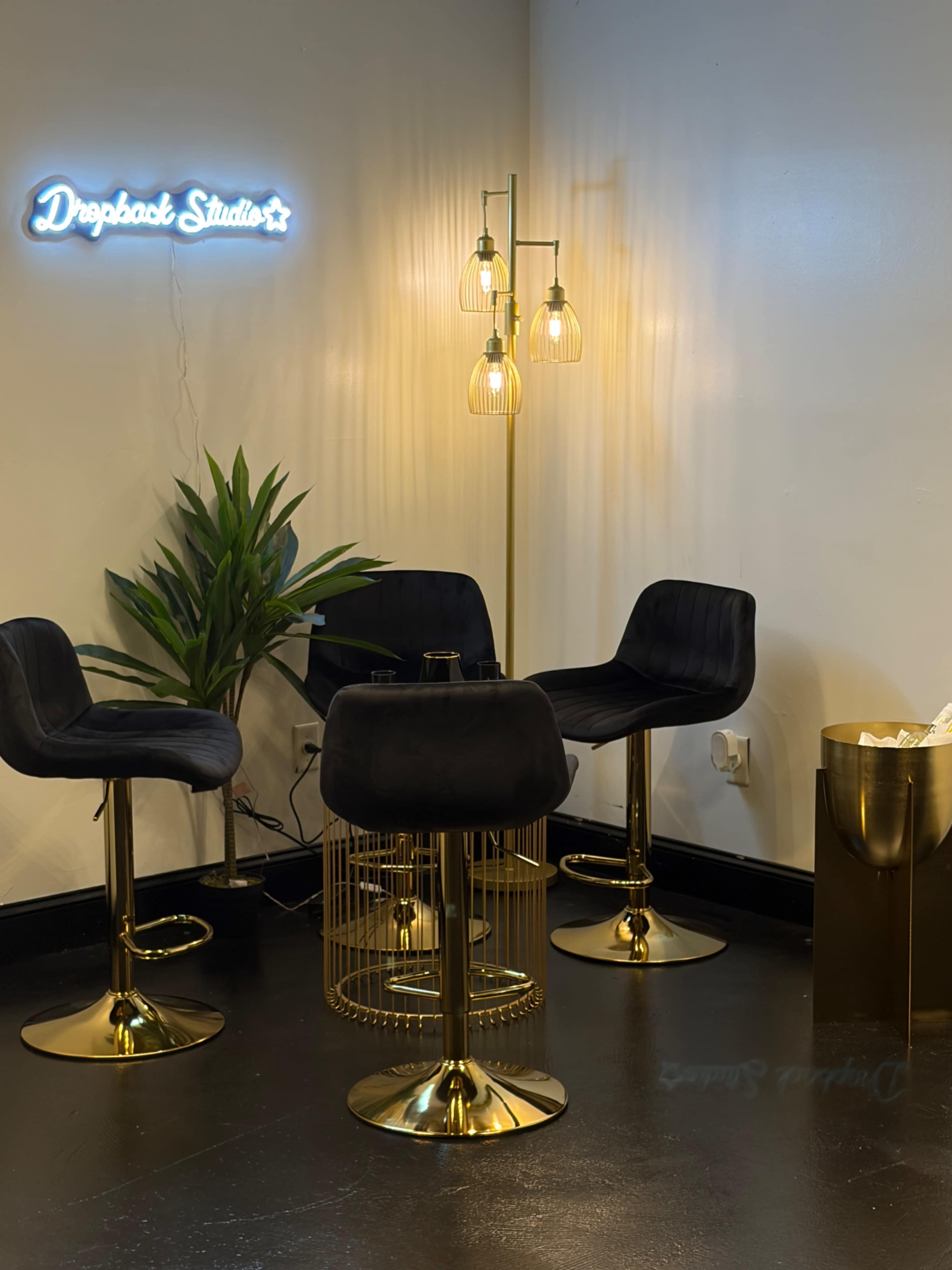 The image shows a modern studio interior featuring black bar stools, a gold floor lamp with hanging bulbs, a potted plant, and a decorative wastebasket, all set against a white wall with a neon sign that reads "Droppook Studio."