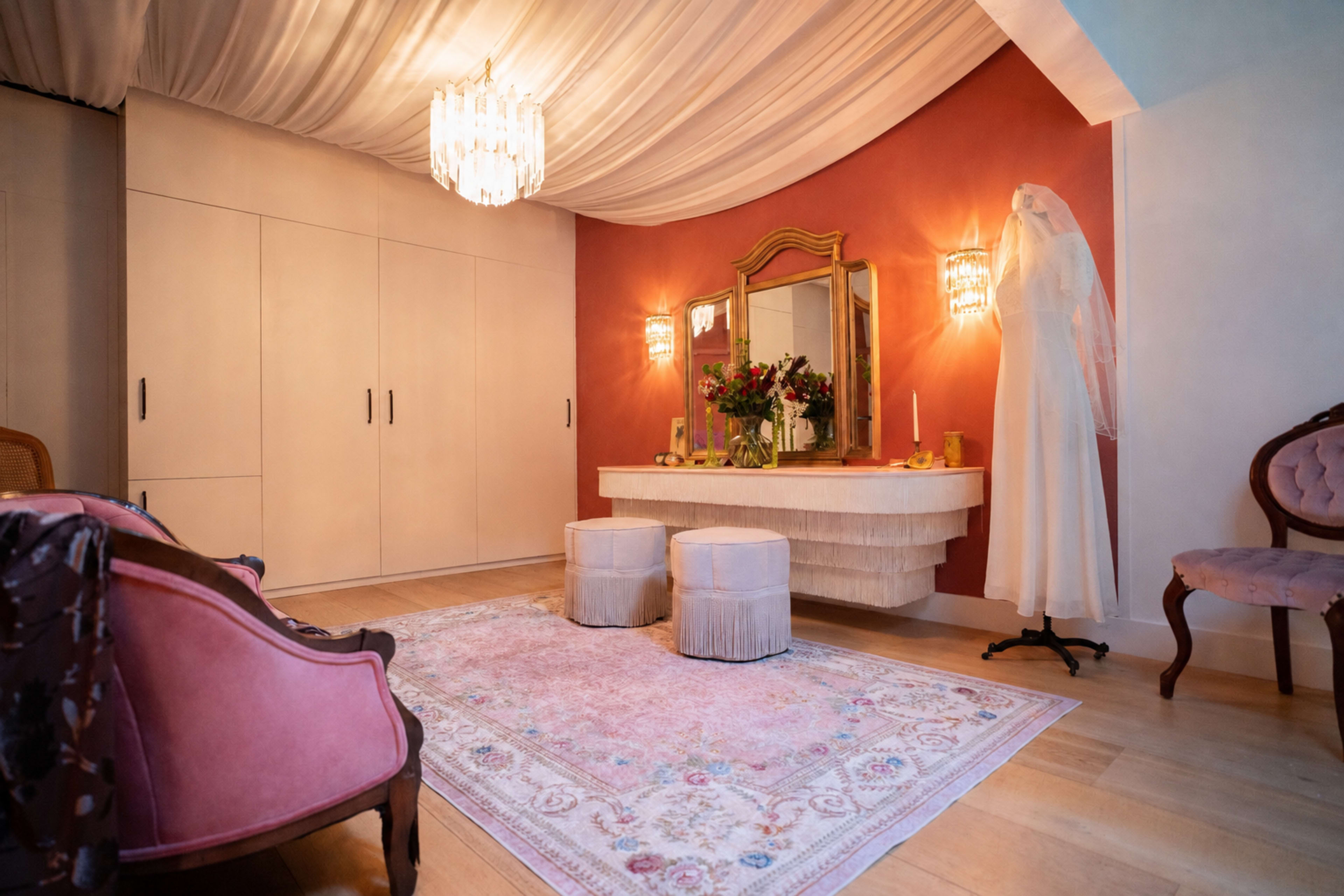 The image shows a well-decorated dressing area with a vintage vanity, two upholstered stools, a wedding dress on a mannequin, and warm lighting under a draped ceiling.