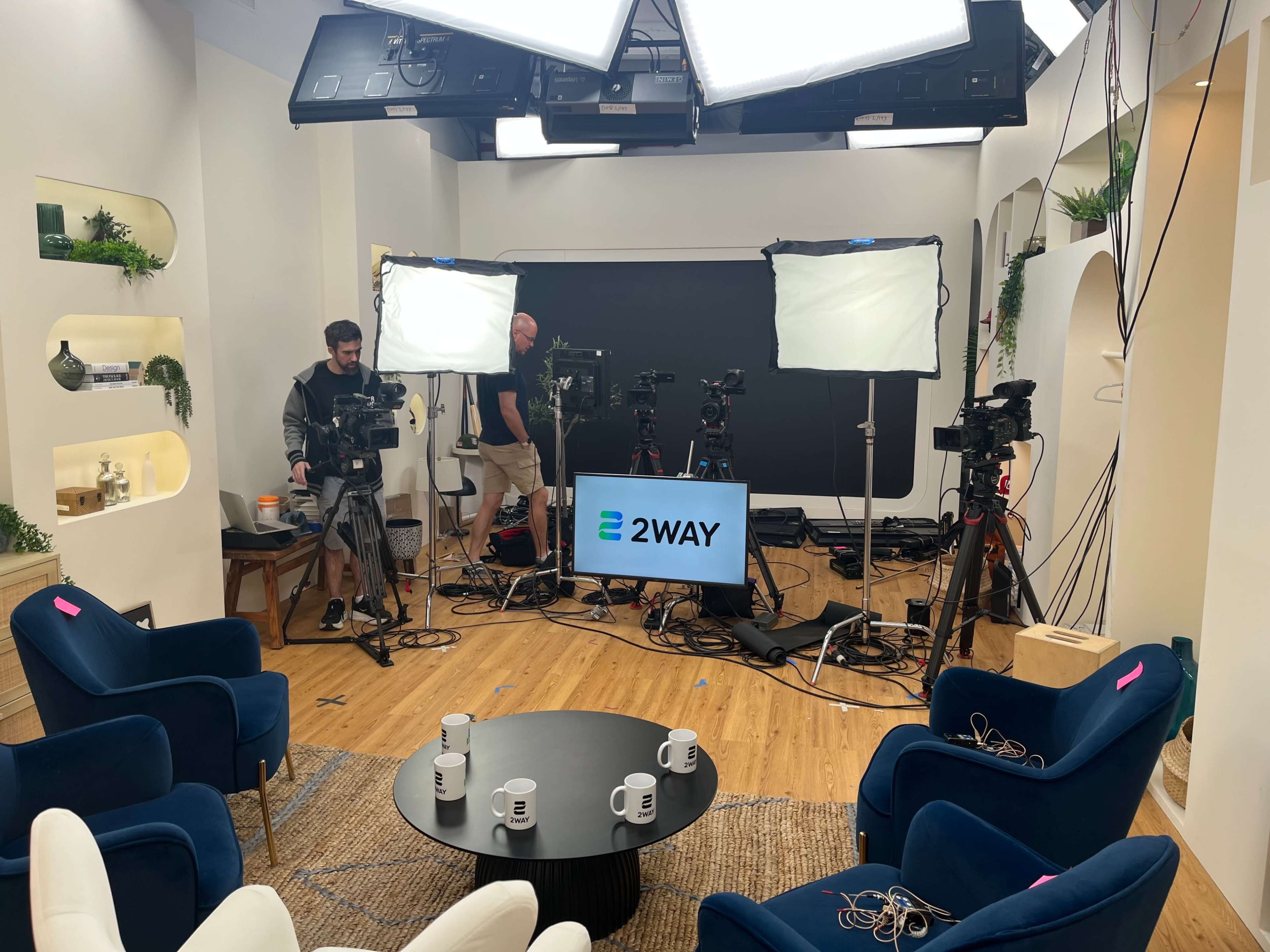 A television studio setup features multiple cameras, lights, and a large display screen with the logo "2WAY," surrounded by blue chairs and a coffee table.