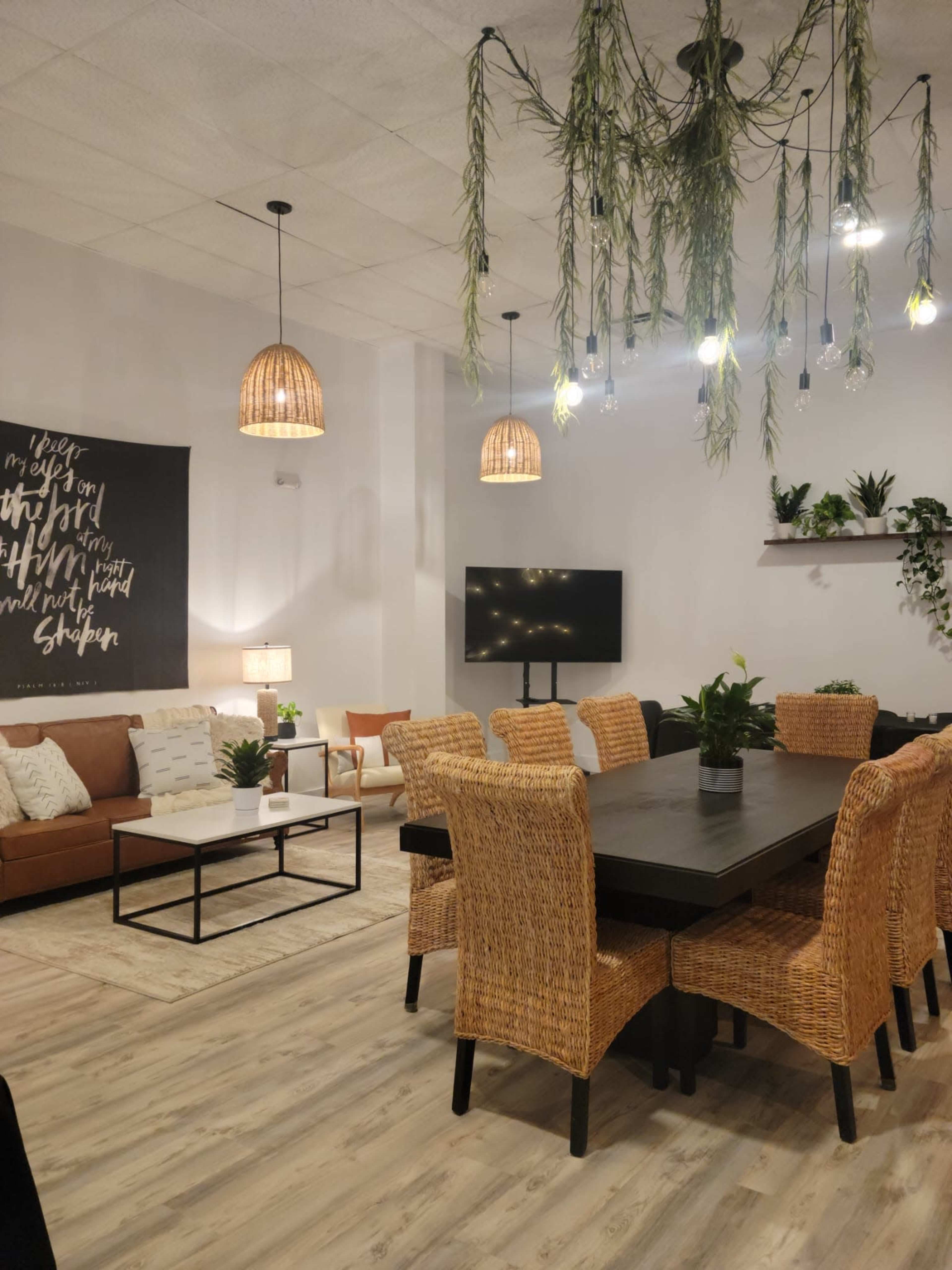 The image shows a modern living space with a large dining table surrounded by woven chairs, a sofa, decorative plants, and pendant lighting.