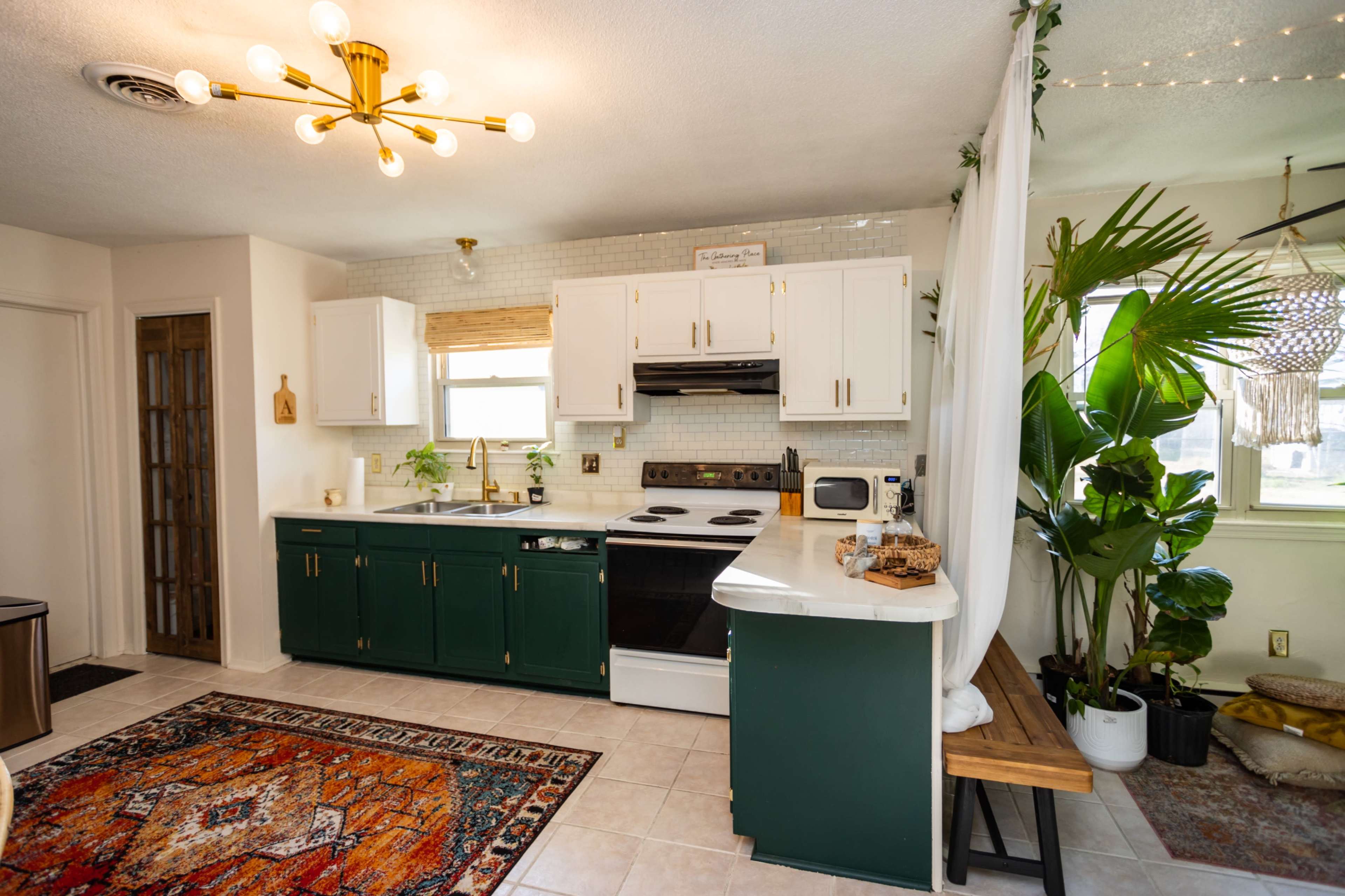 The kitchen features green cabinetry, a white countertop, and a seating area with a bench, accompanied by a large potted plant and a colorful area rug.