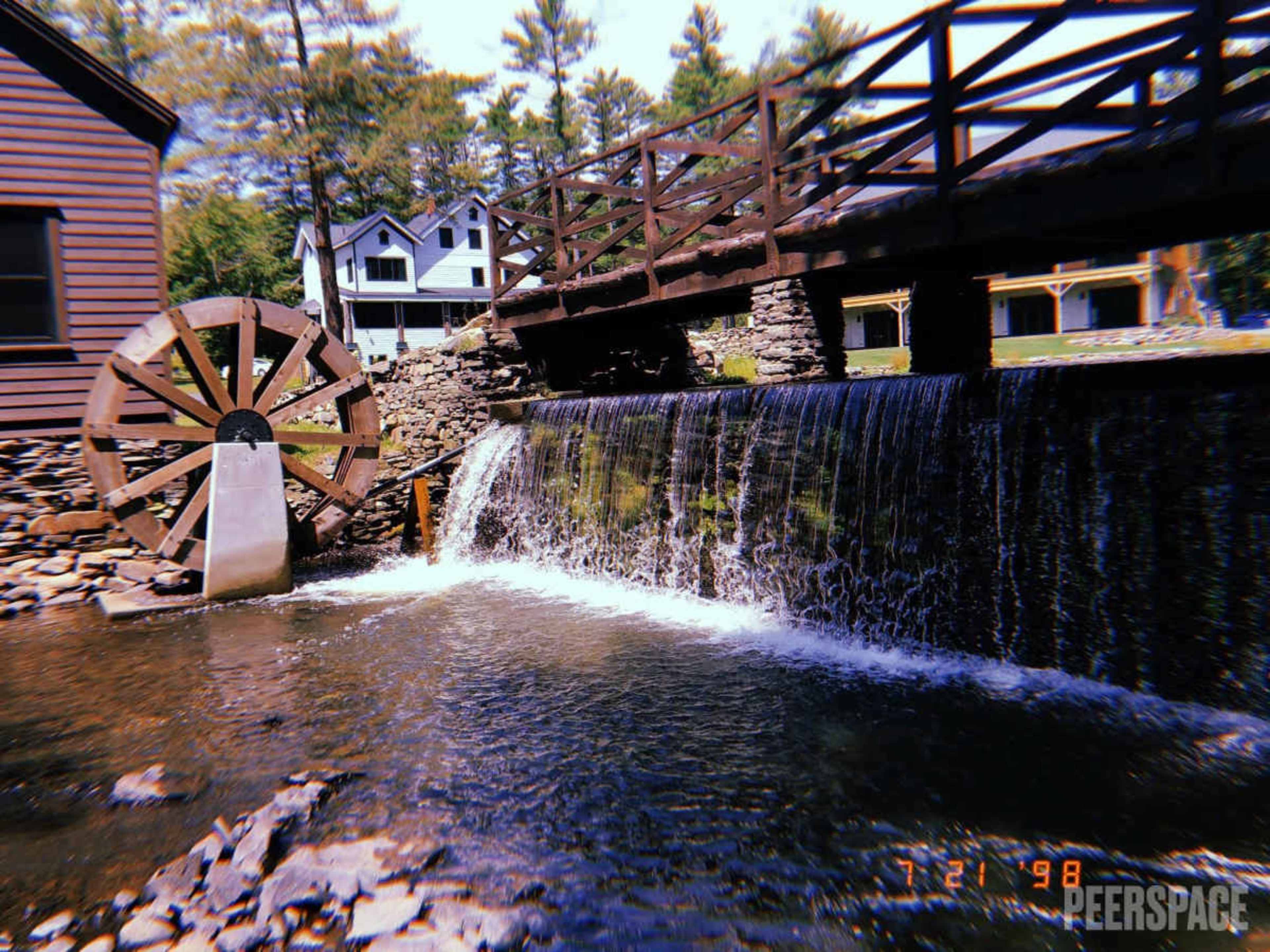 The image shows a waterwheel beside a small waterfall, with a wooden bridge crossing over the water and houses visible in the background.