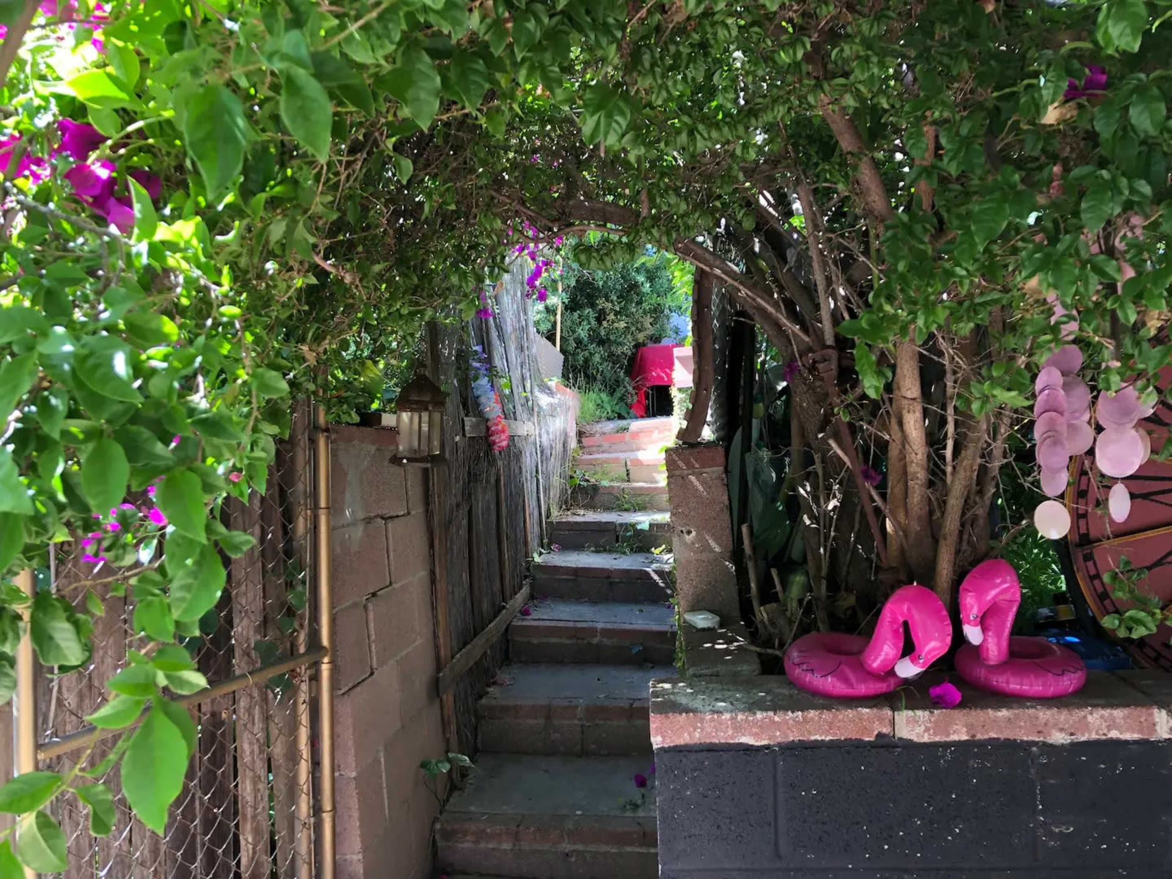 A stone pathway leads through a dense, green archway with colorful flowers on either side, leading to a small red table in the distance.