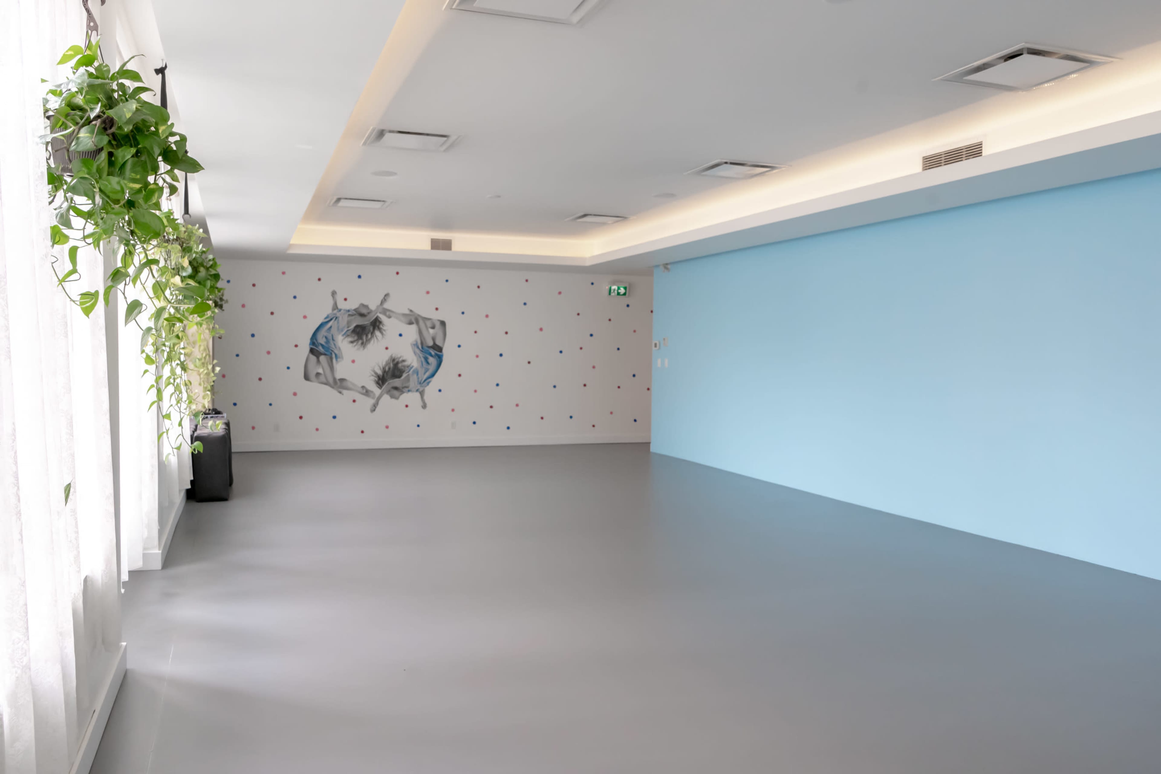 A spacious, empty room features a light blue wall with a colorful dotted pattern and a mural, alongside large windows adorned with indoor plants.