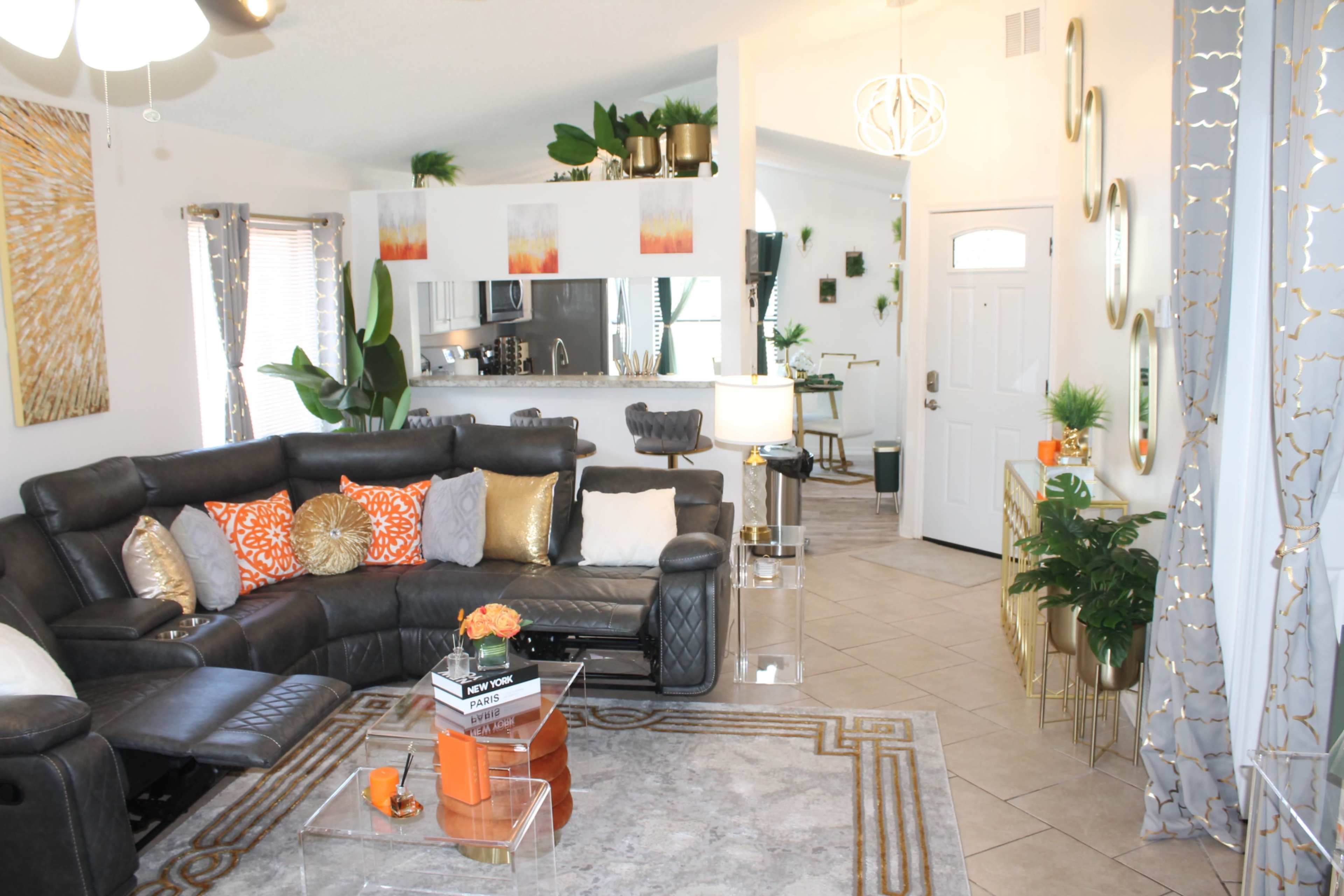 A modern living room with a sectional sofa, colorful cushions, a glass coffee table, and a doorway leading to a kitchen area adorned with plants and decorative accents.