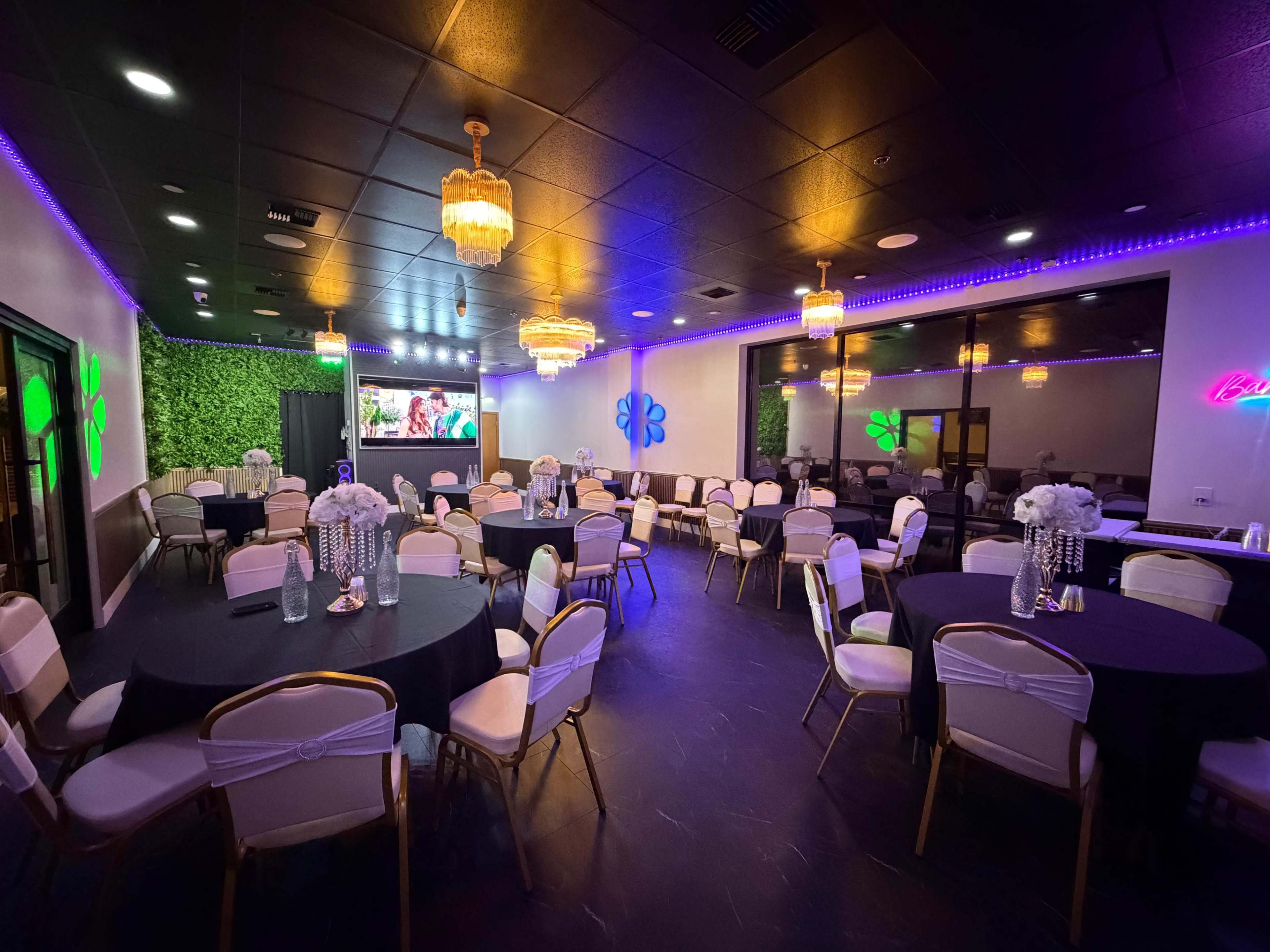The image shows an elegantly arranged event space with round tables covered in black cloth, white chairs, and decorative floral centerpieces, illuminated by purple and gold lights.