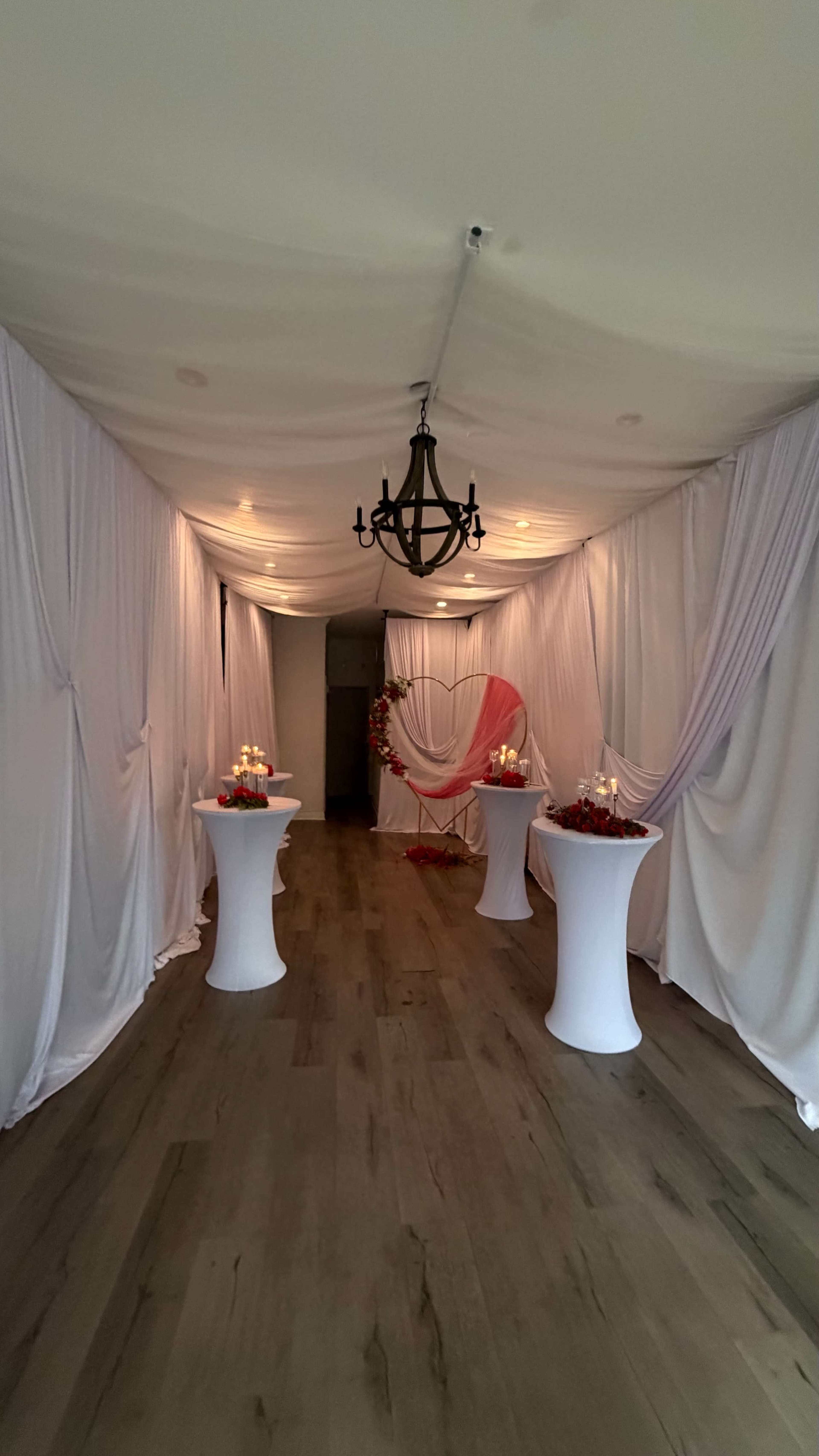 The image shows a decorated indoor hallway with draped fabric, several white cocktail tables, and a chandelier.