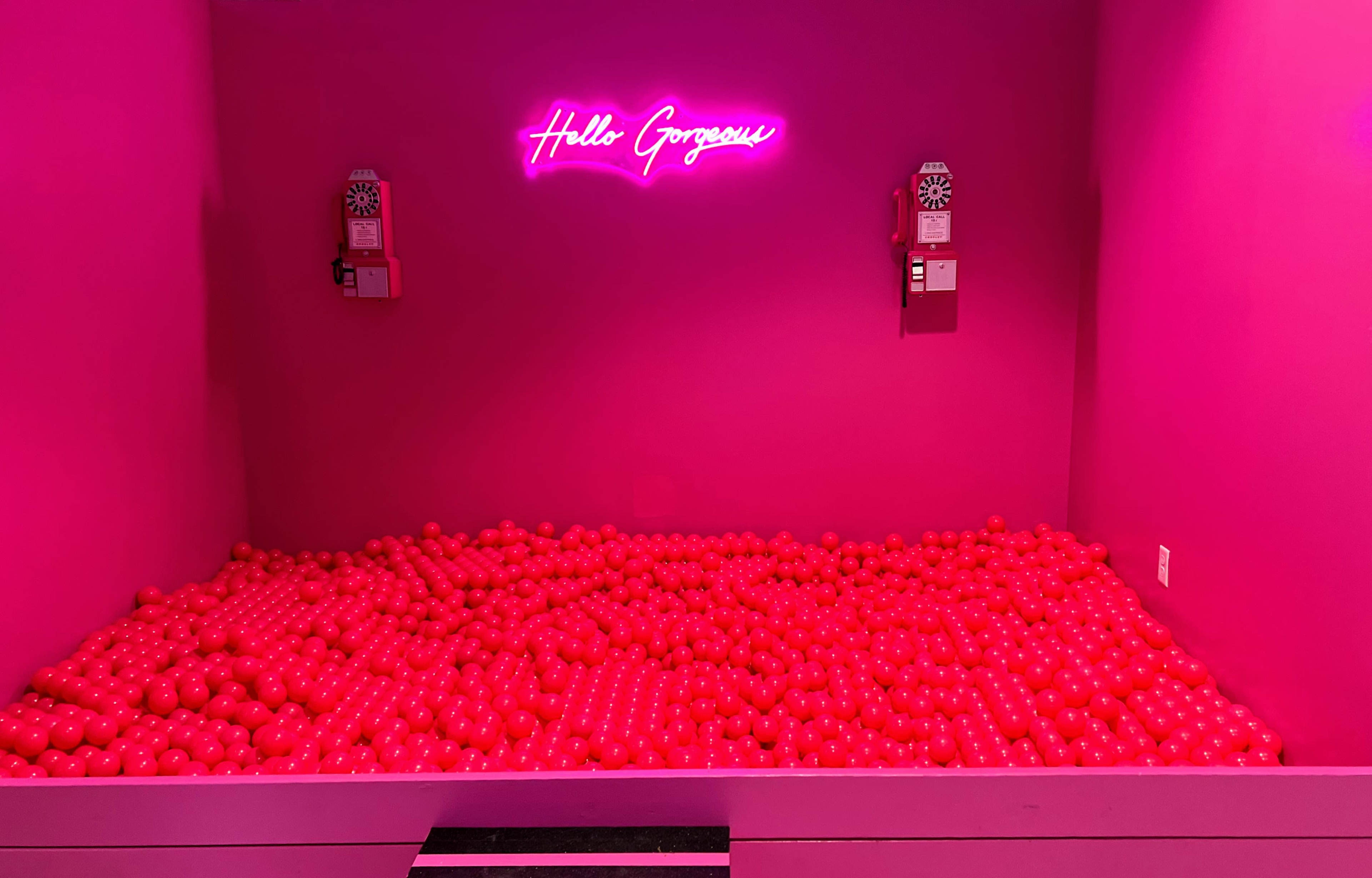 A room filled with red plastic balls, illuminated by a pink neon sign that reads "Hello Gorgeous."