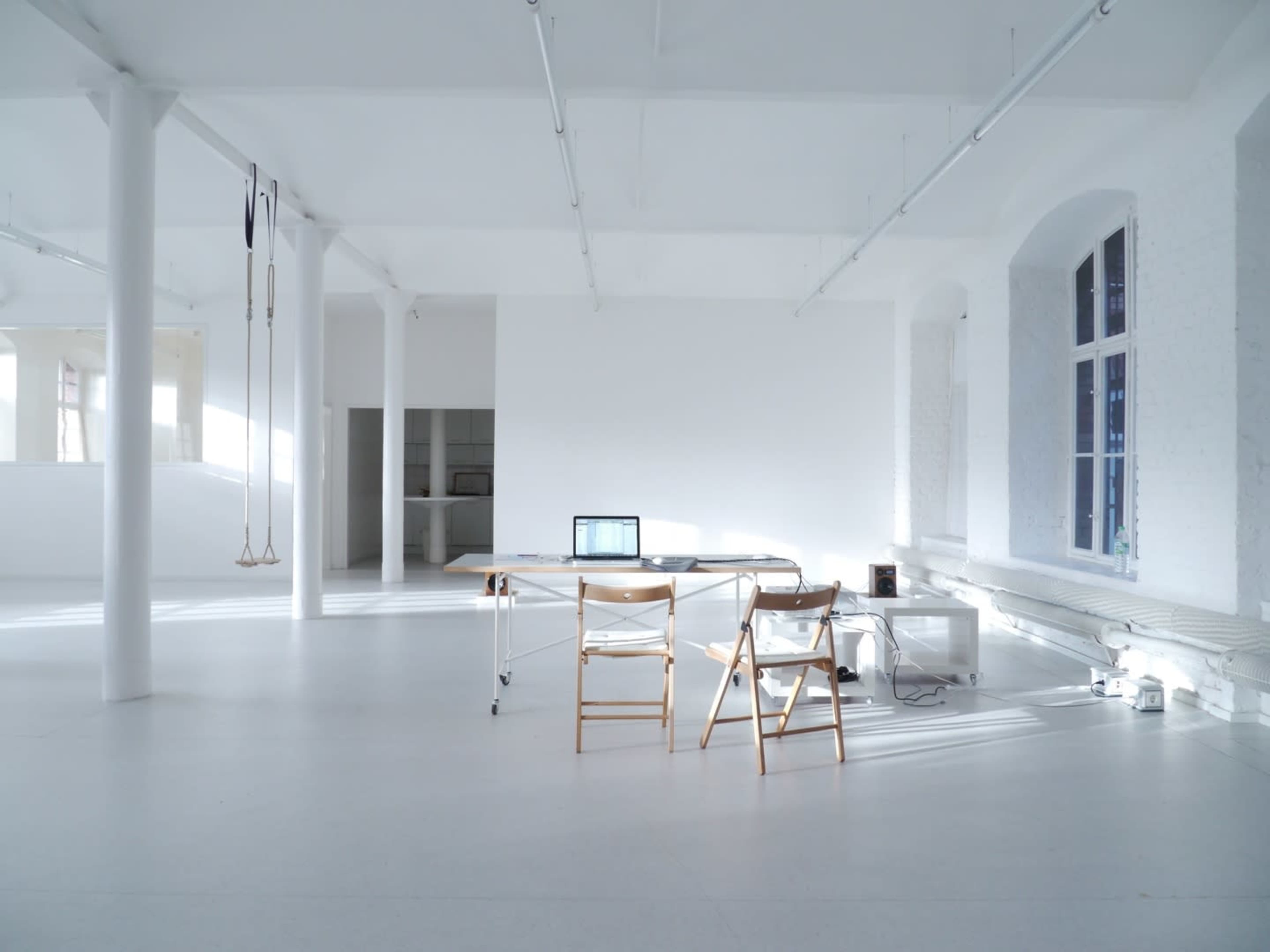A large, minimalistic room with white walls and floors features a simple desk, two chairs, and a laptop, illuminated by natural light from windows.