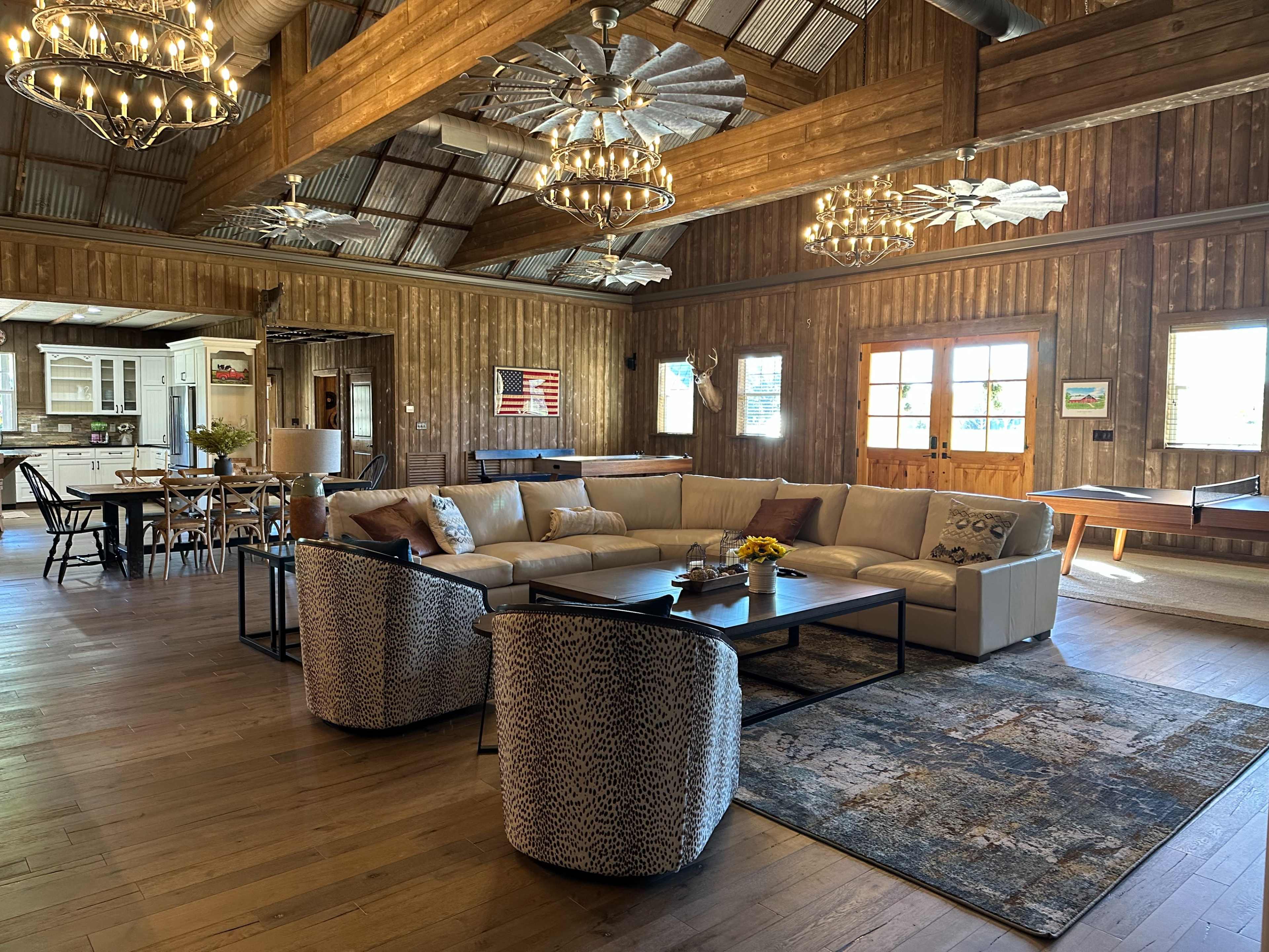The spacious living area features a large sectional sofa, two patterned armchairs, a coffee table, and rustic wood walls illuminated by hanging light fixtures.