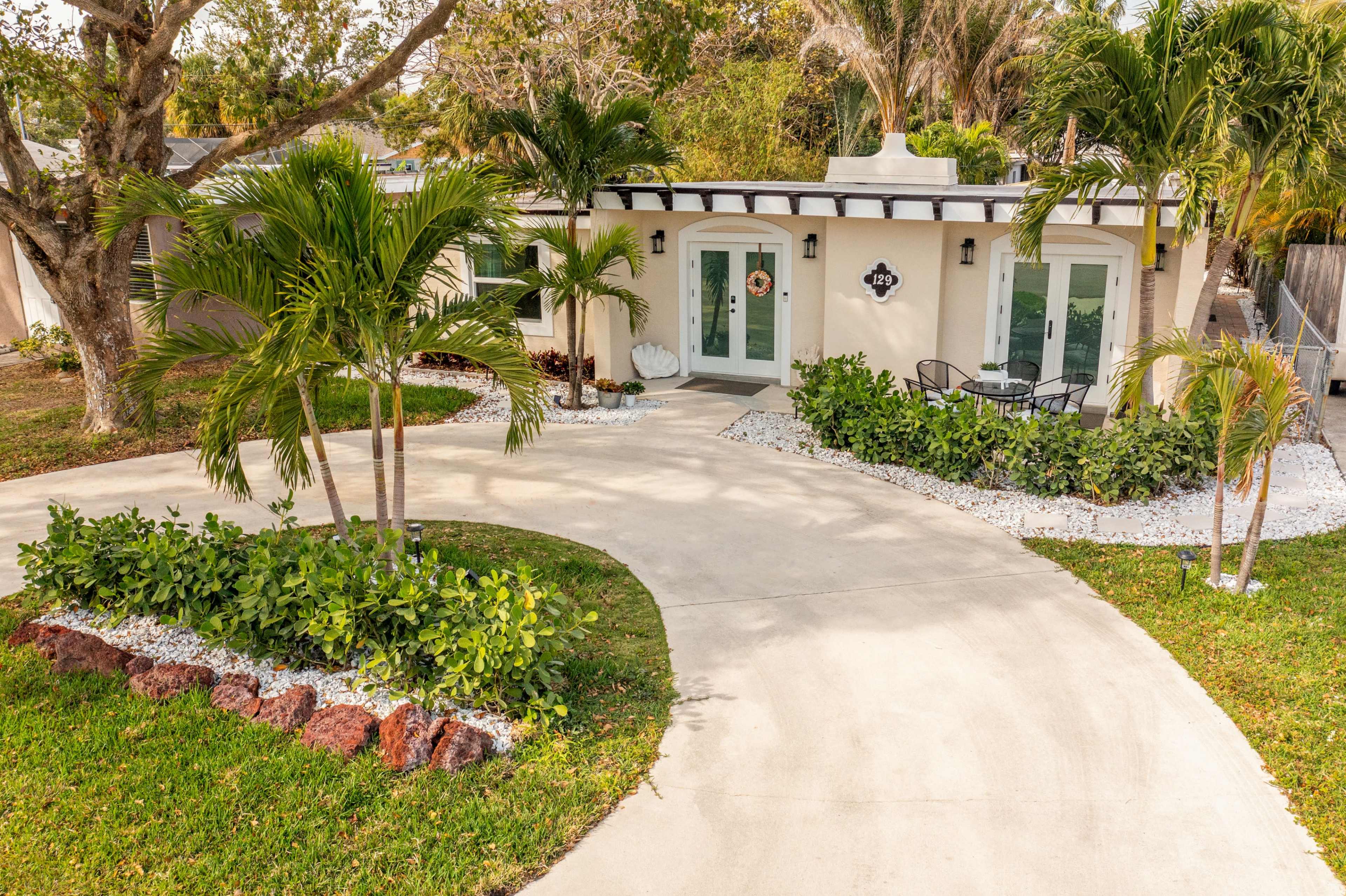 The image shows a well-landscaped house with a circular driveway, bordered by palm trees and decorative stones.