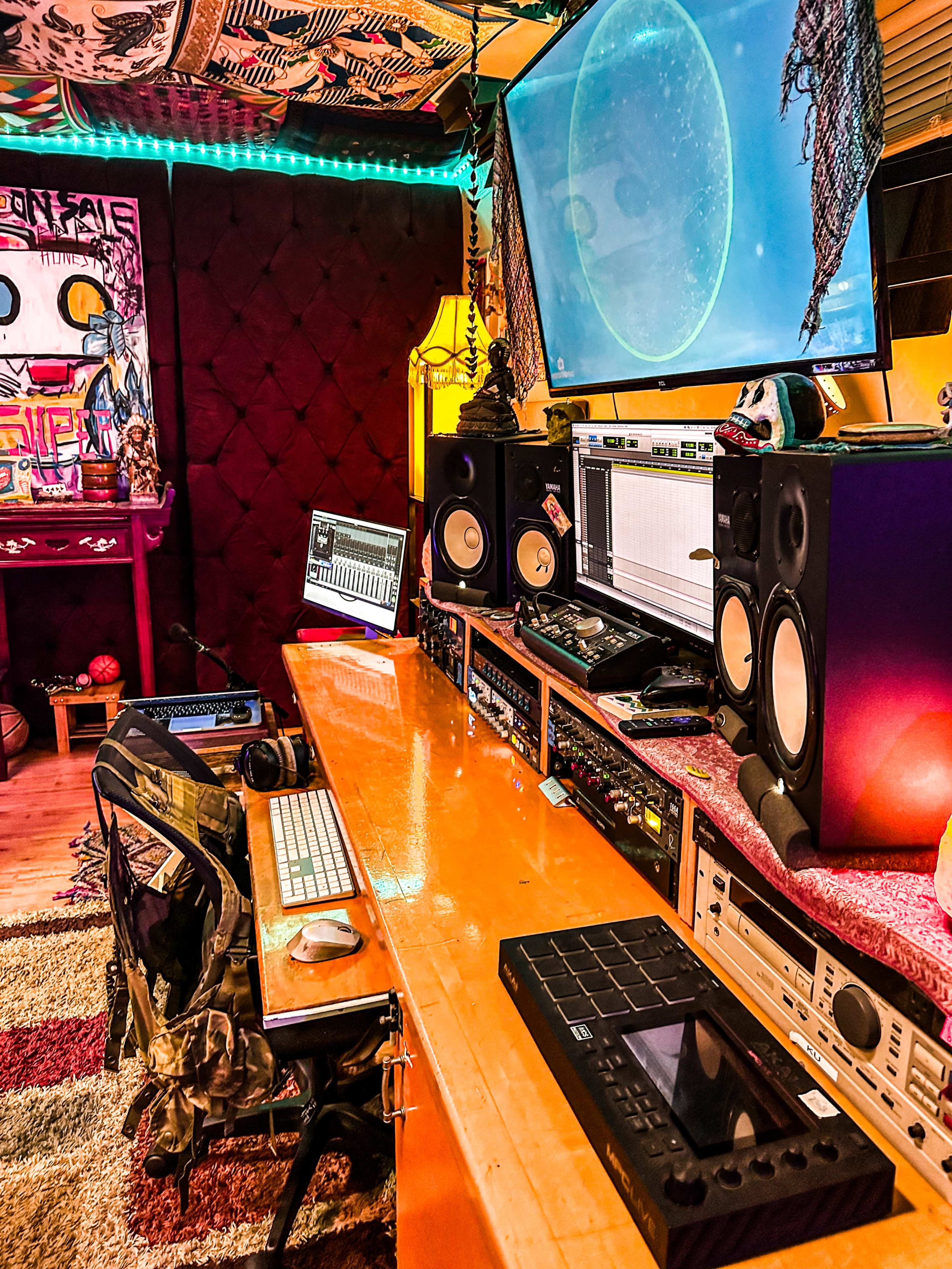 A music production studio features a long desk with computers, sound equipment, speakers, and a large monitor displaying a visual.