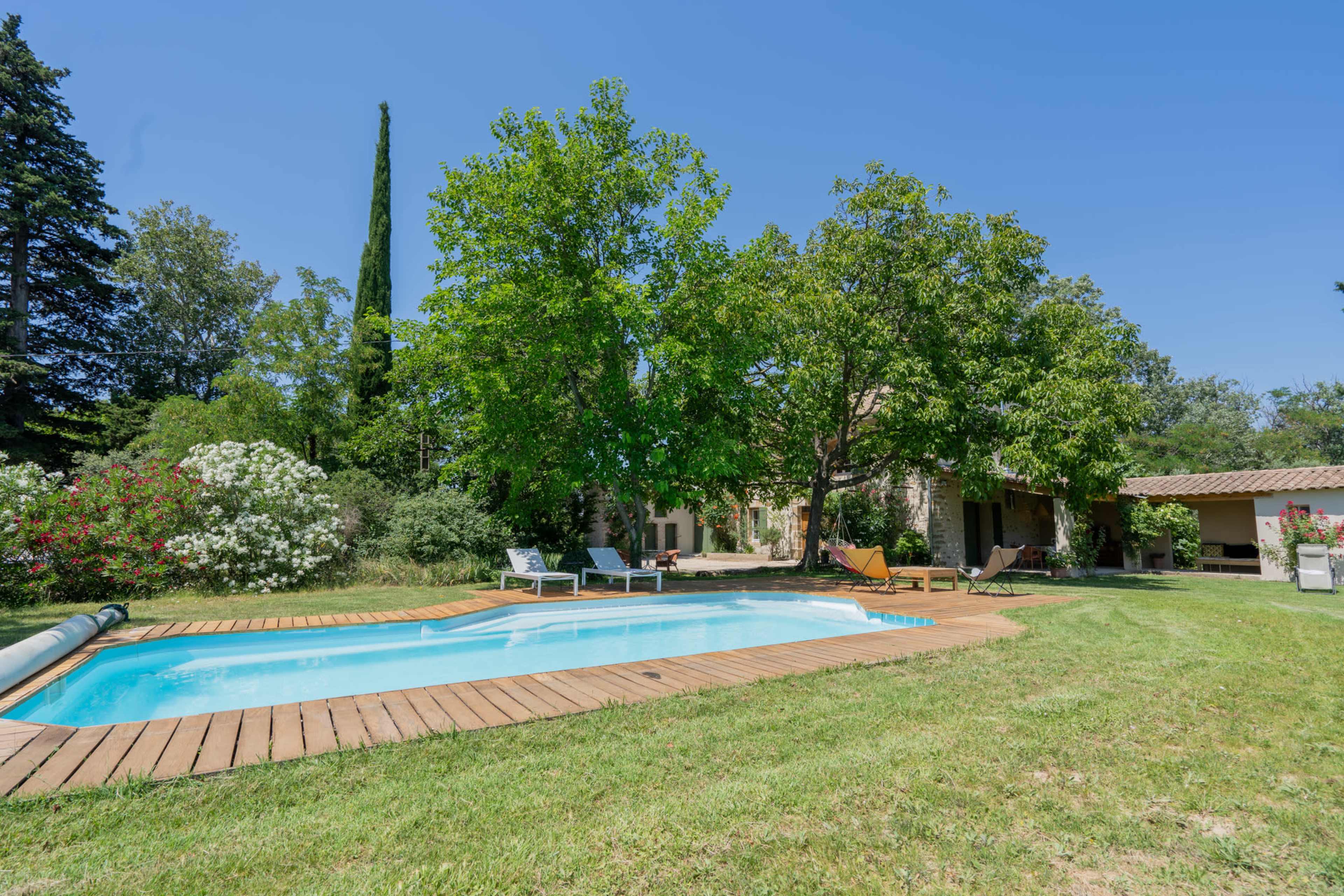 The image shows a backyard with a swimming pool surrounded by wooden decking, lush greenery, and a house partially obscured by trees.