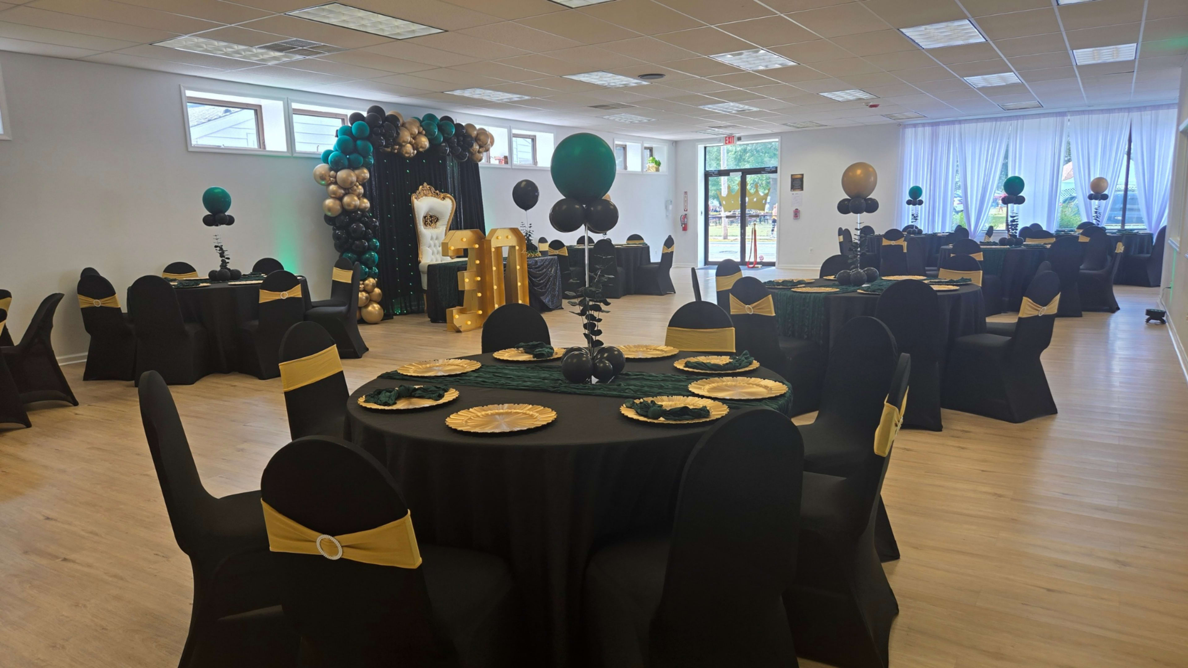 The image shows a large event space set up with black round tables, gold plates, and decor featuring green, black, and gold balloons.
