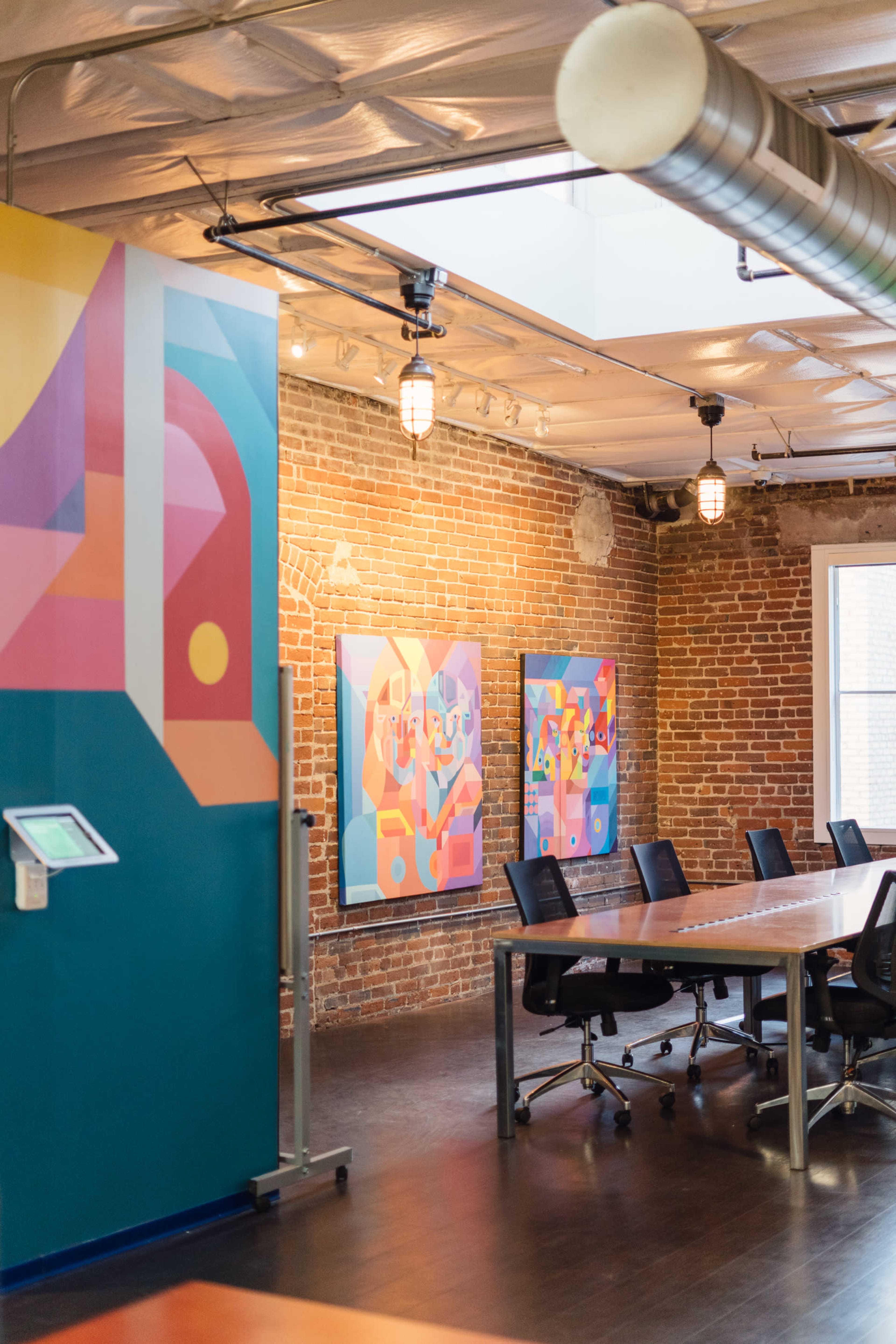 A modern conference room featuring exposed brick walls, colorful abstract artwork, and a large wooden table surrounded by black chairs.